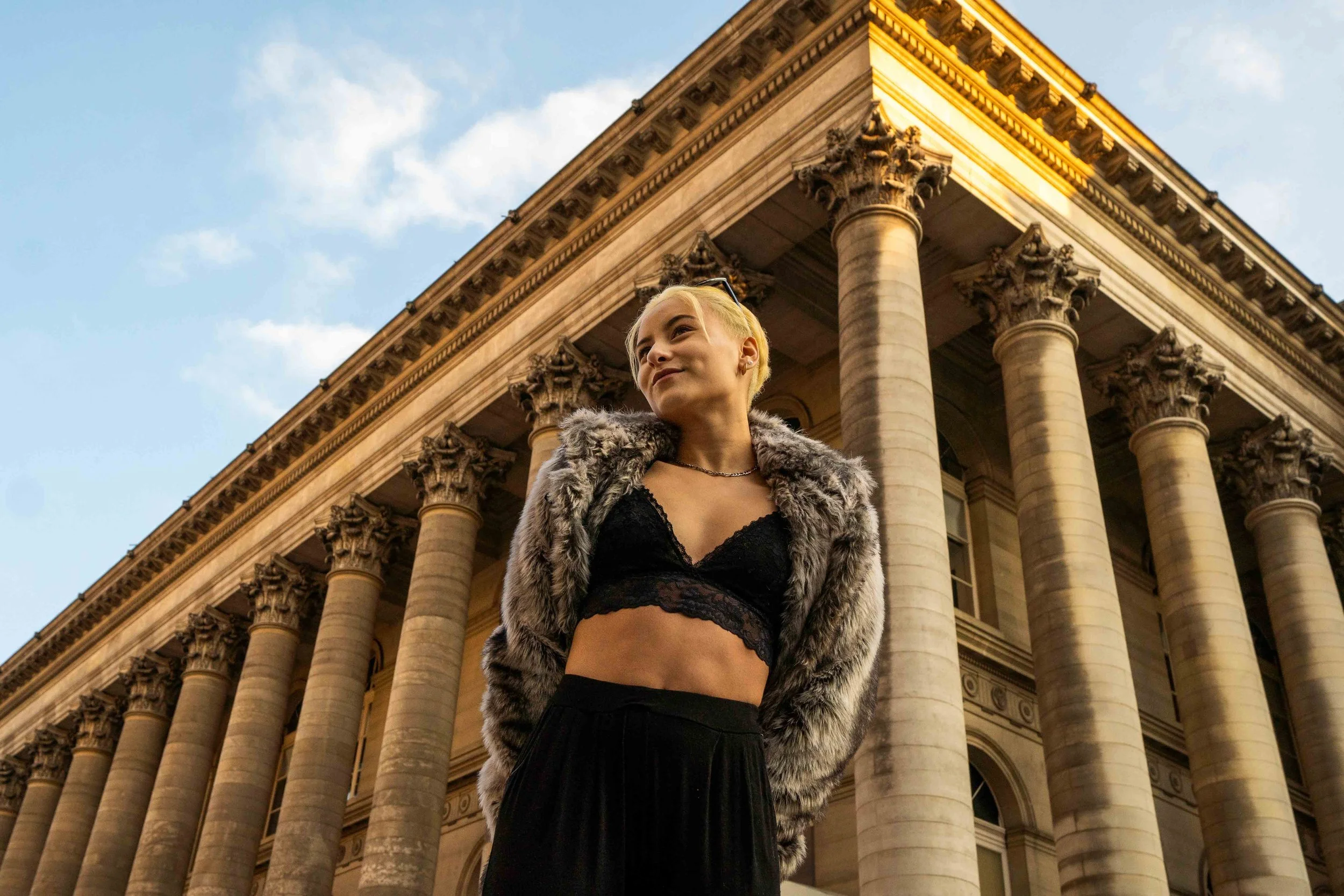 A young woman with short blonde hair standing in front of a historic building with large columns, wearing a black lace crop top, black skirt, and a gray fur coat, looking off to the side with a confident expression.
