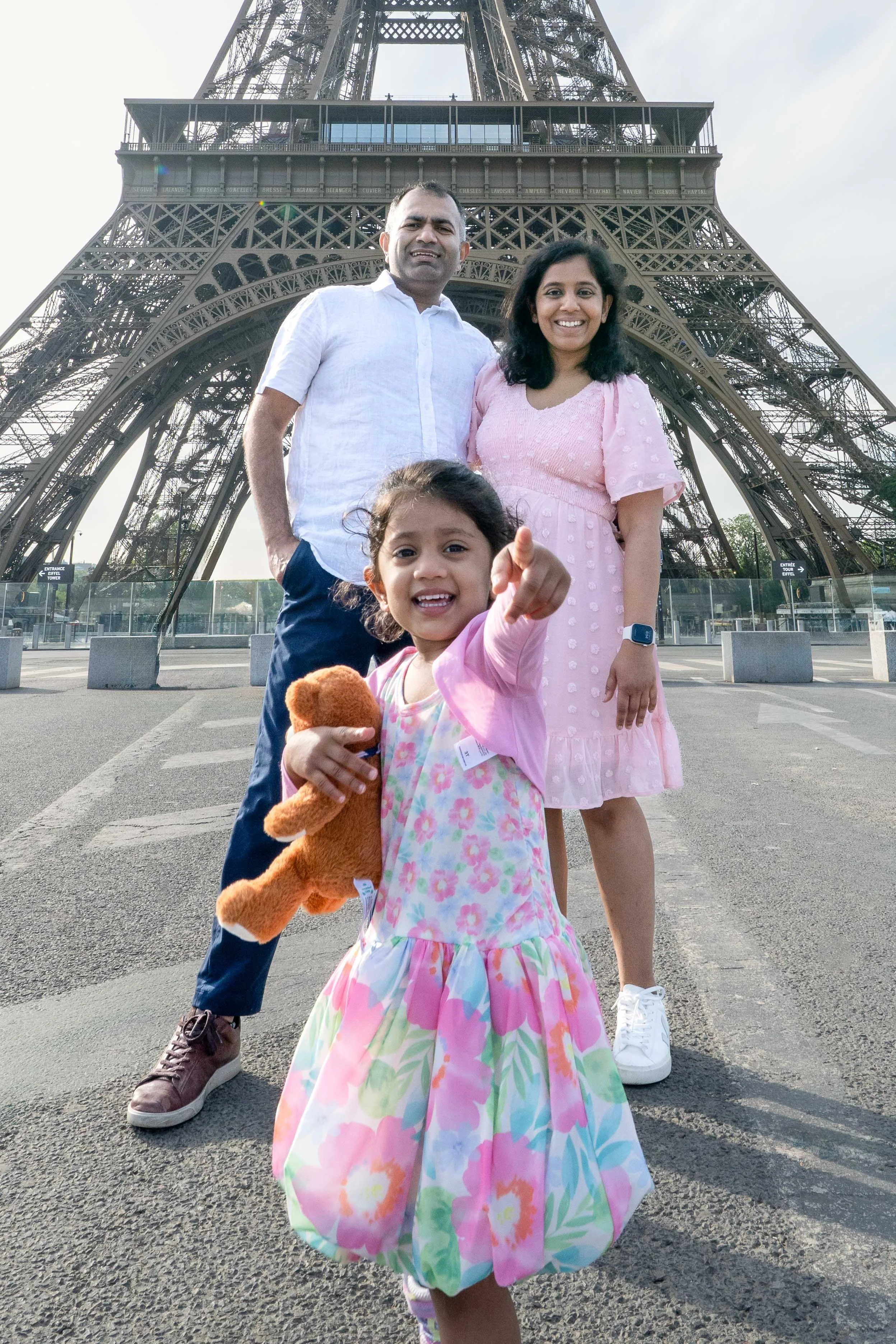 A family of three standing in front of the Eiffel Tower during daytime. The young girl in the foreground is smiling and pointing at the camera, holding a teddy bear. The man and woman are behind her, smiling, with the Eiffel Tower as the backdrop.