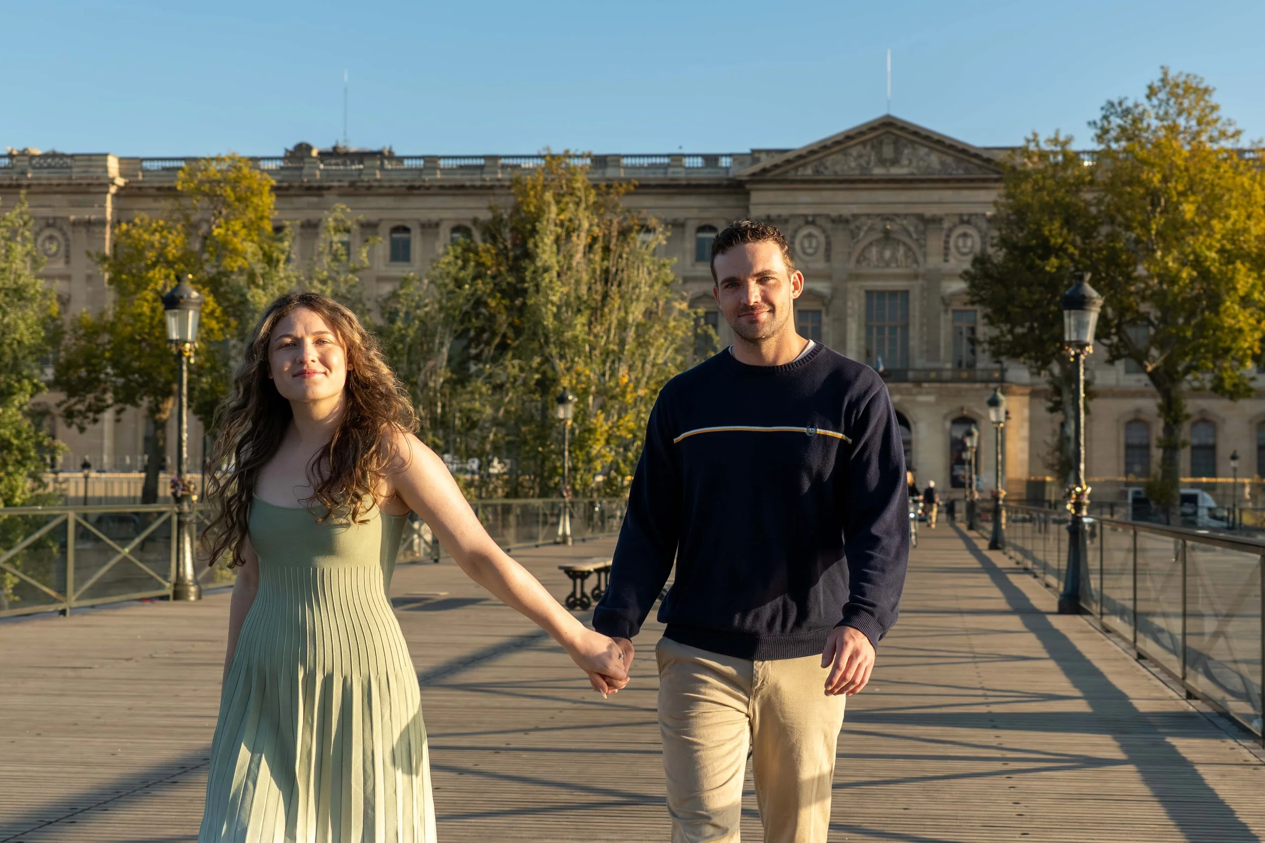 A young couple holding hands and walking on a wooden bridge in front of a historic building surrounded by trees during daytime.