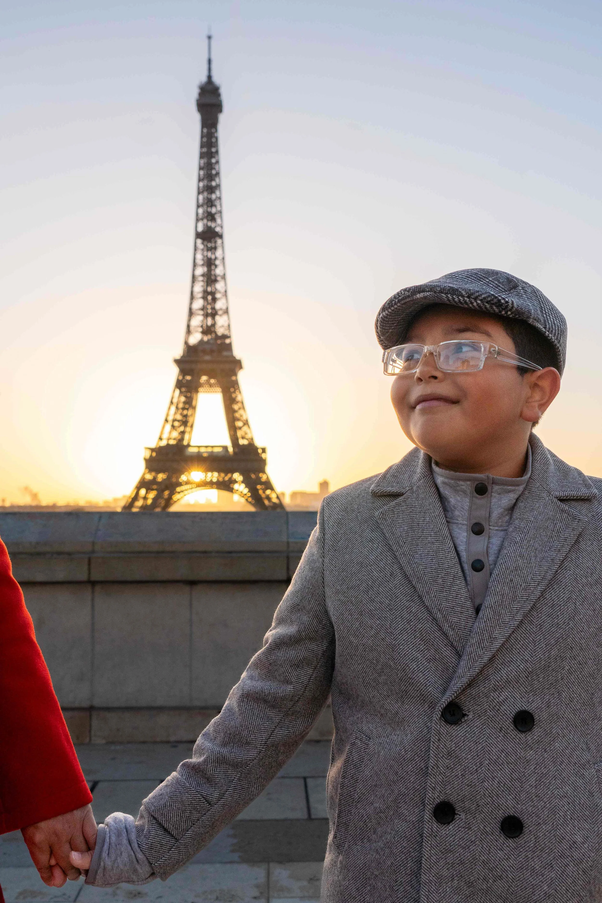 A young boy wearing glasses, a gray cap, and a gray coat holding hands with someone during sunset in front of the Eiffel Tower in Paris, France.