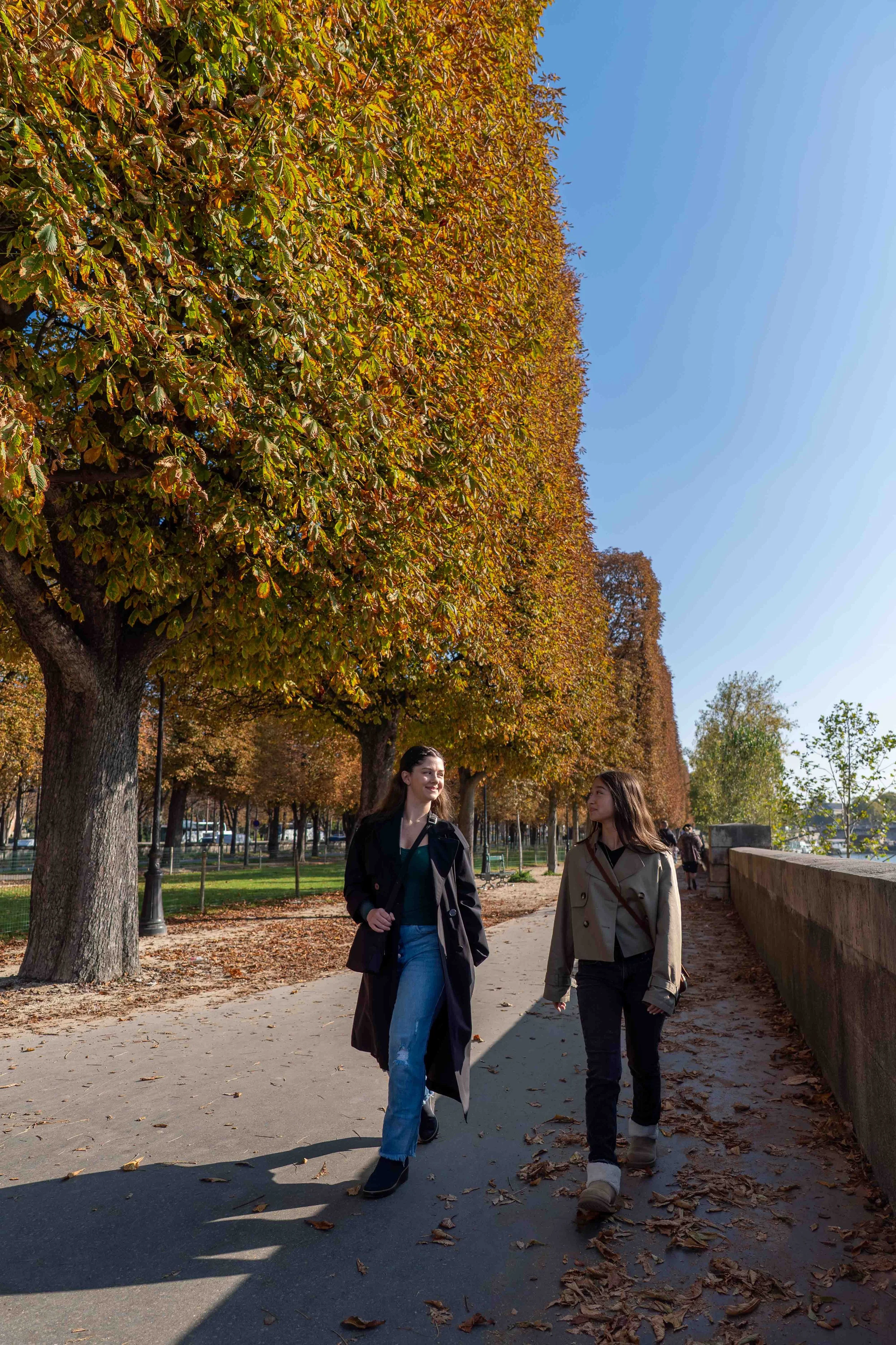 Two women walking along a paved pathway in a park, surrounded by tall trees with autumn-colored leaves, under a clear blue sky.