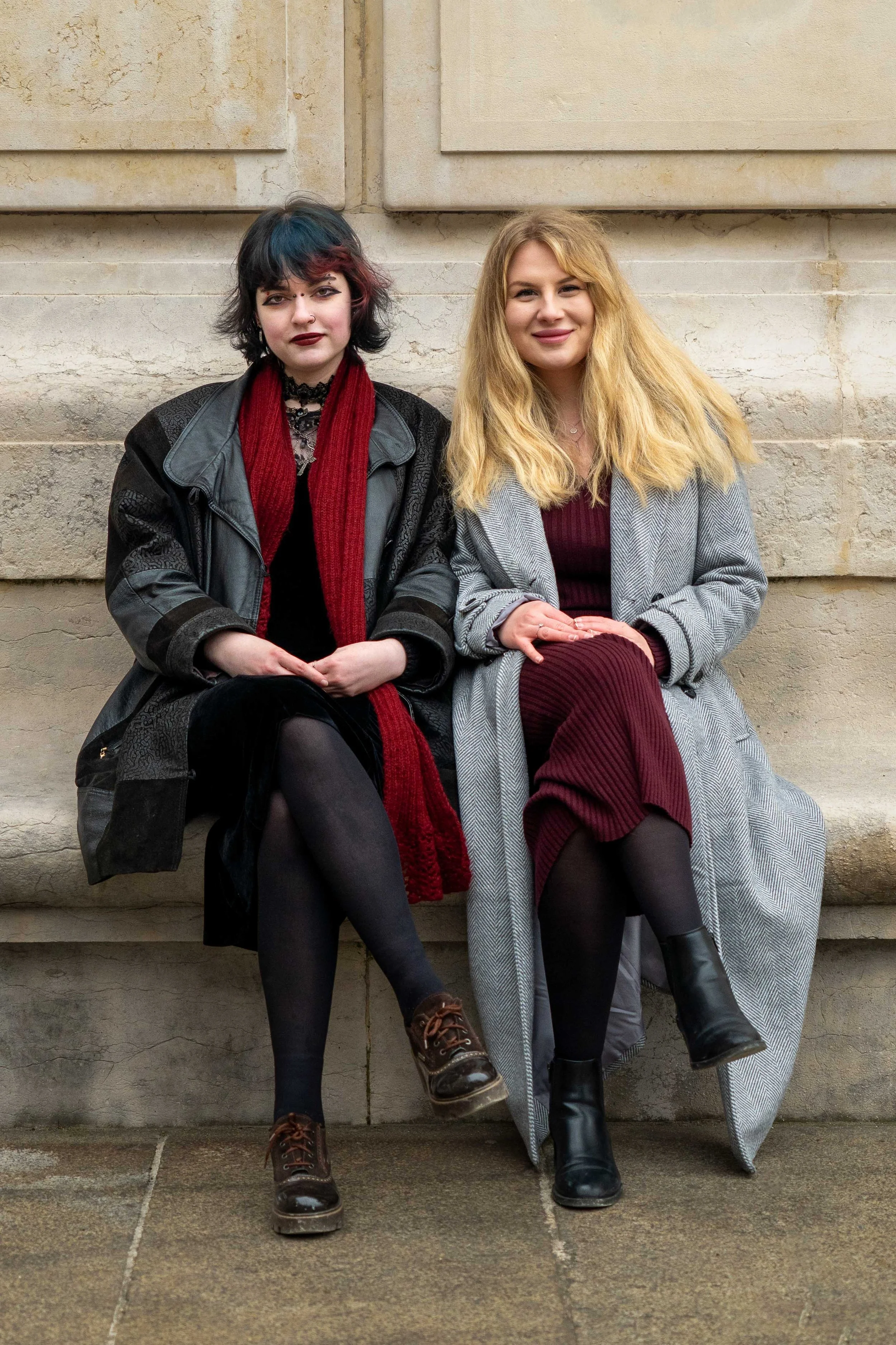 Two women sitting on a stone bench against a beige stone wall, smiling, wearing coats and black tights.
