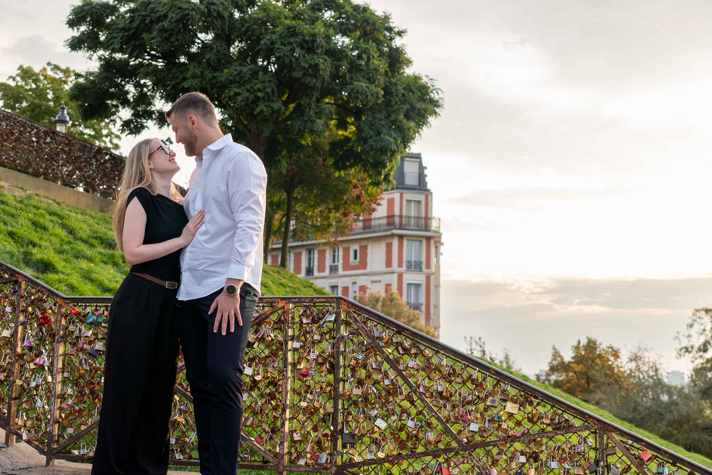 A couple standing close to each other, smiling and about to kiss, outdoors near a bridge with love locks, a grassy hill, and trees with a building in the background.