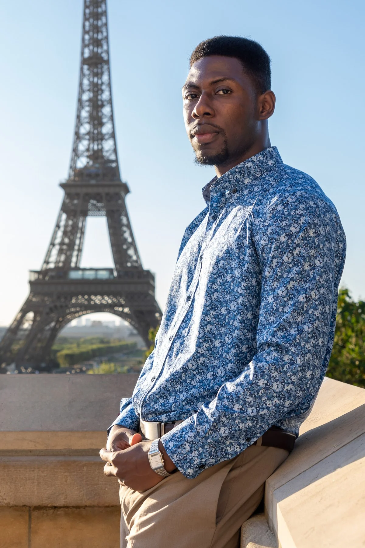 A man in a blue floral shirt and beige pants with a silver watch is standing outdoors in front of the Eiffel Tower in Paris, France, during daylight.