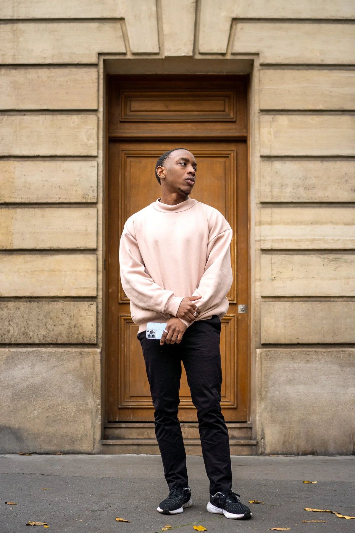A young man stands on a city sidewalk in front of a large wooden door set in a stone building, holding a smartphone in his hand, wearing a beige sweatshirt, black pants, and sneakers, looking to his right.