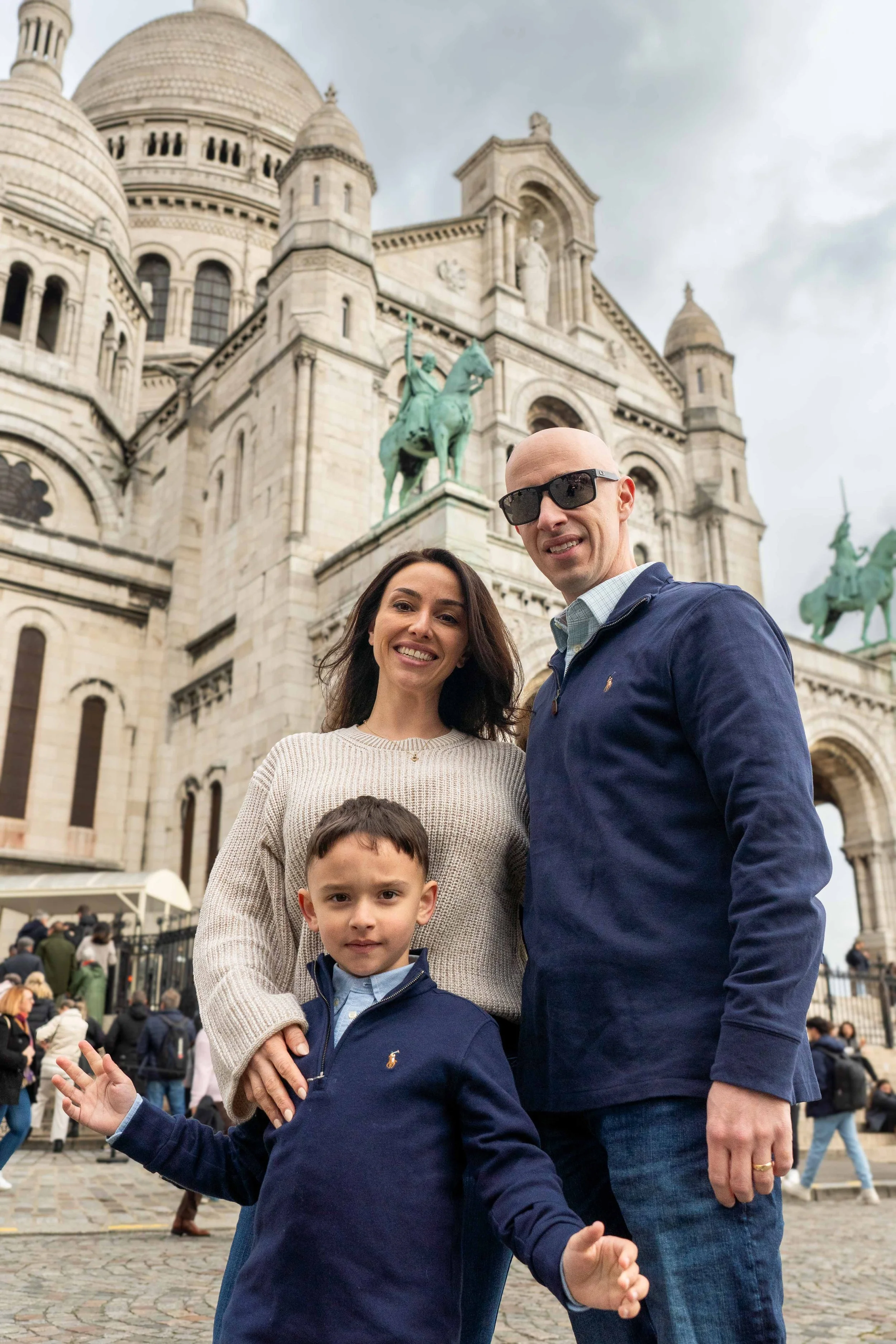A family of three posing in front of the Sacré-Cœur Basilica in Paris, with a cloudy sky overhead. The family includes a woman with long dark hair, a man with sunglasses and a shaved head, and a young boy in a navy blue sweater.