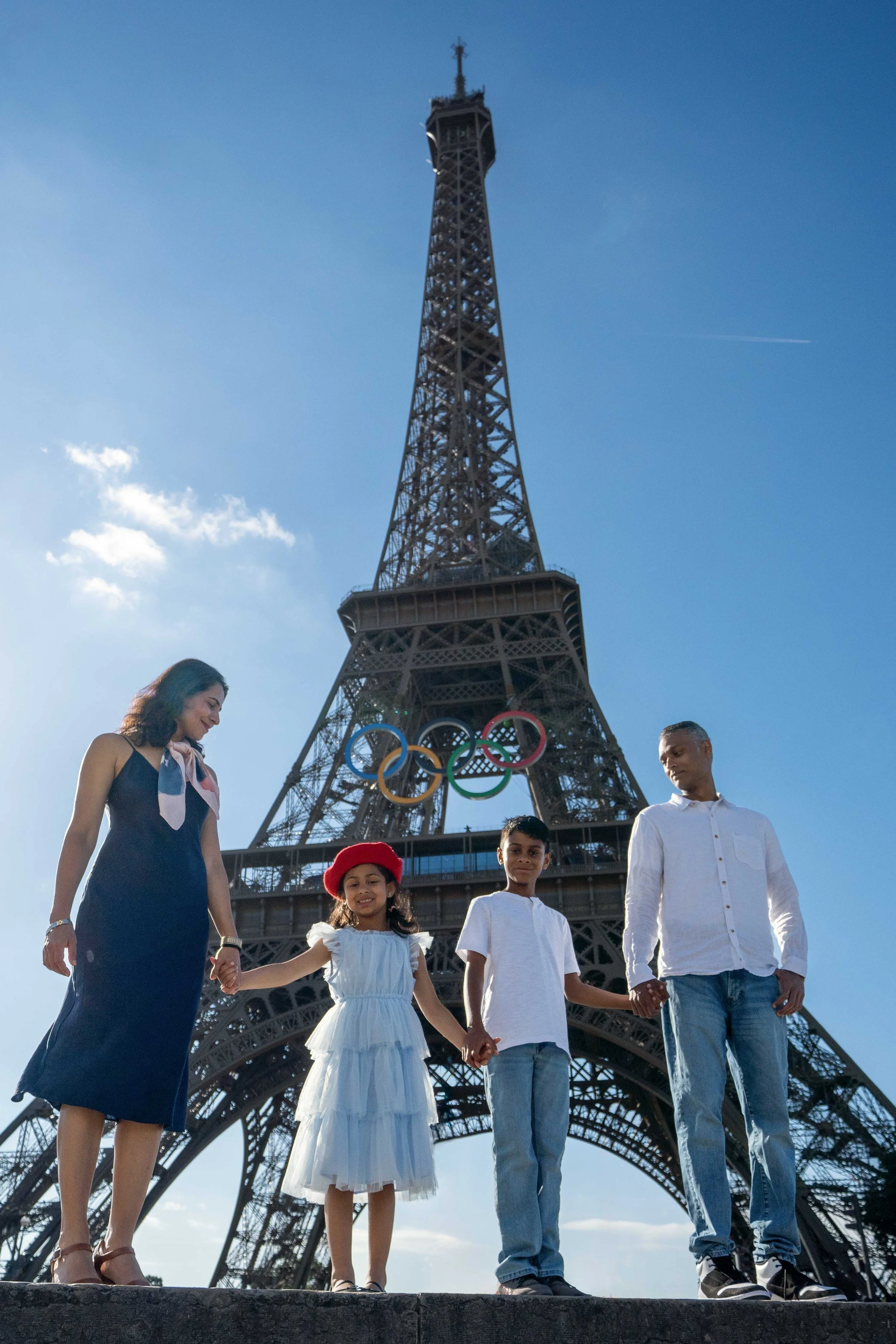 A family of four holding hands in front of the Eiffel Tower with Olympic rings hanging on it, under a clear blue sky.