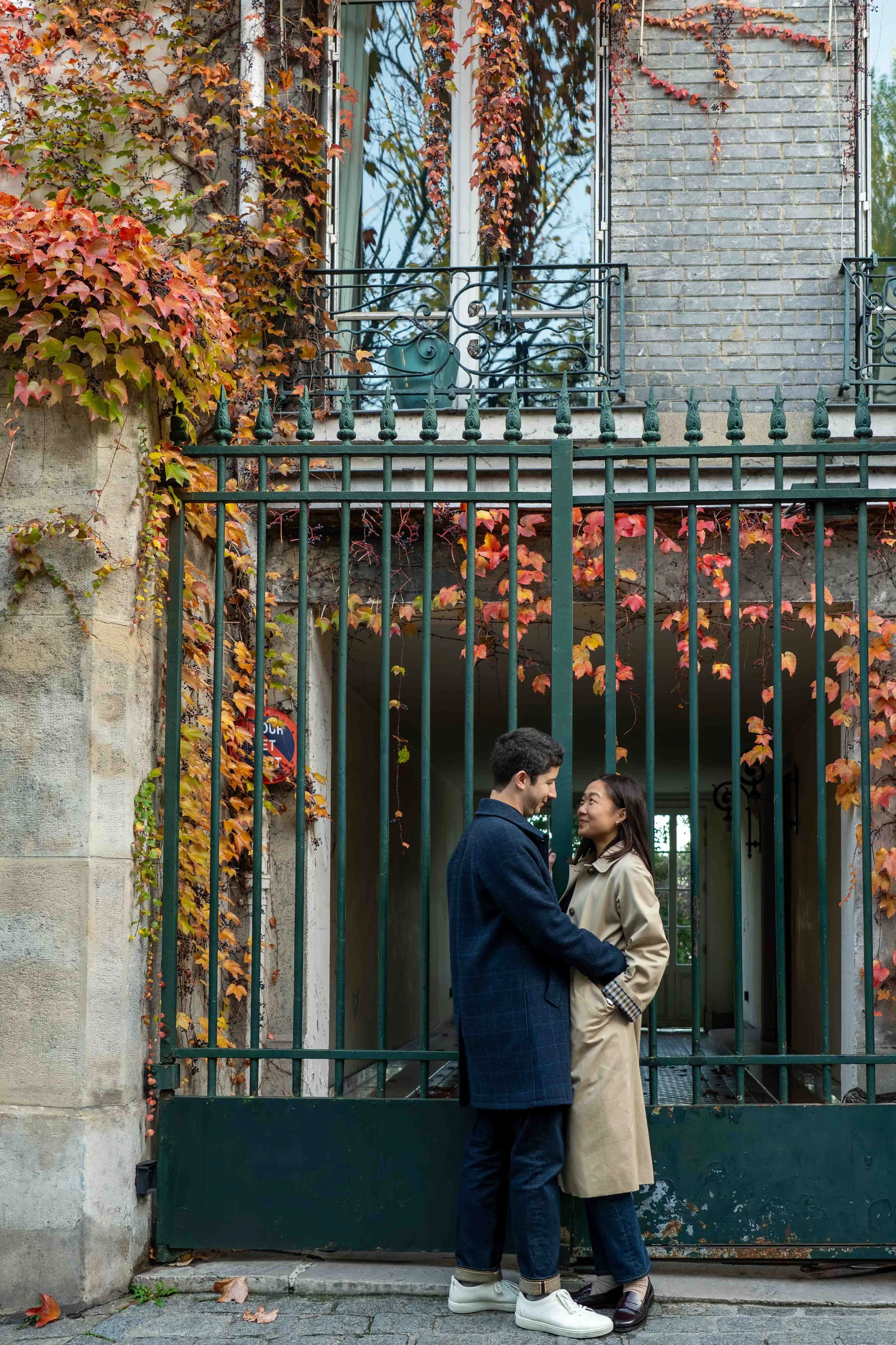 A man and woman standing close together behind a green iron gate, looking into each other's eyes, with a building and autumn leaves in the background.