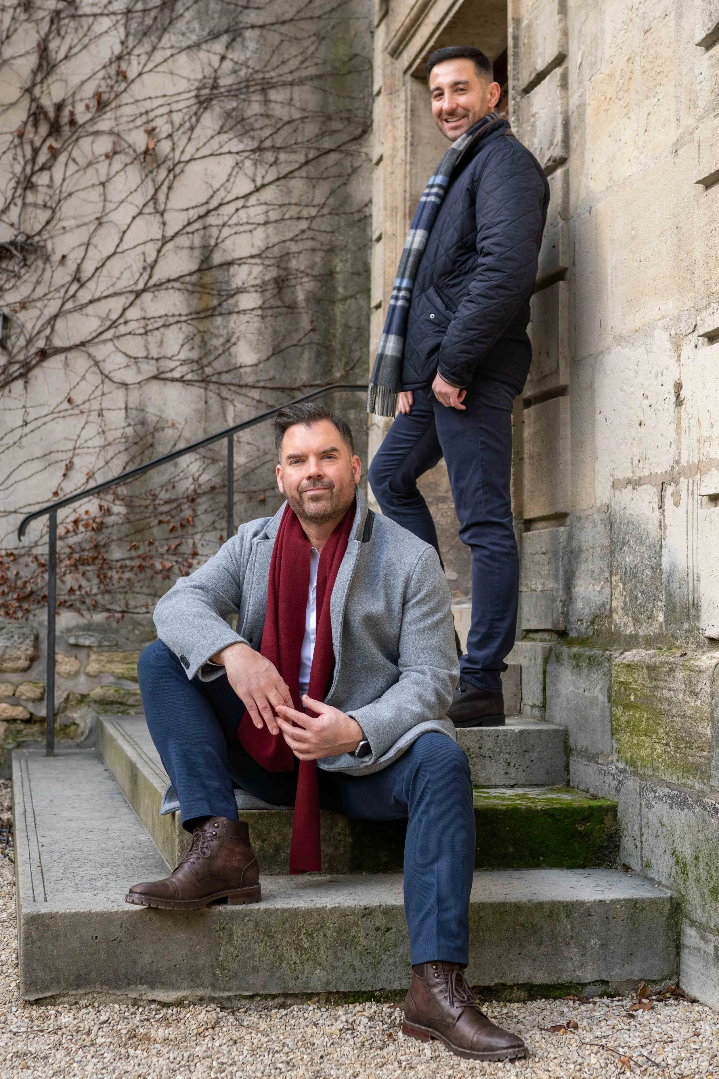 Two men are sitting on steps outside, one standing behind the other. The man sitting in front has dark hair, a beard, and is wearing a gray coat, maroon scarf, dark pants, and brown boots. The man standing behind has short dark hair, a beard, and is 