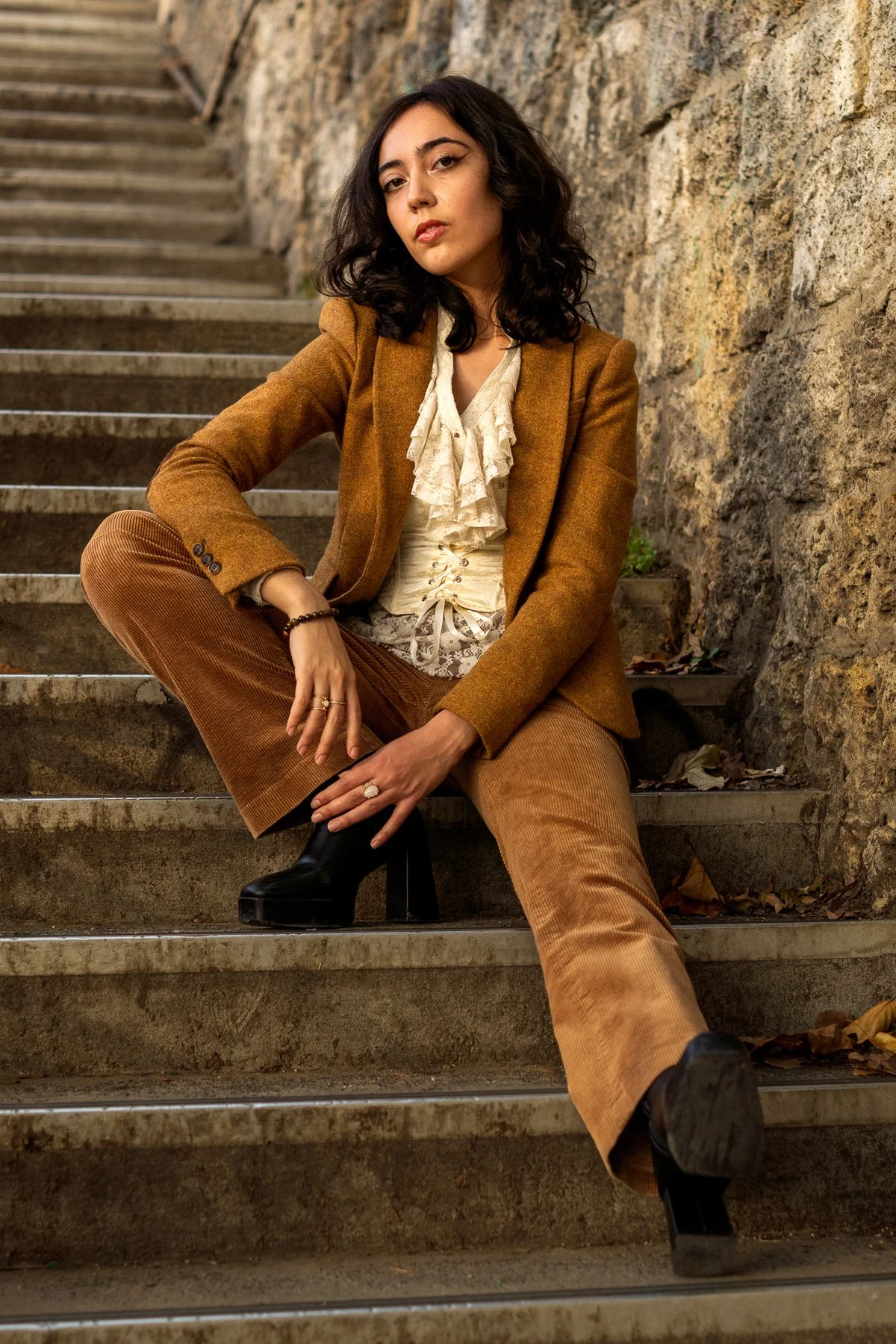 A woman with black curly hair sitting on outdoor stone stairs, wearing a brown blazer, light-colored ruffled blouse, brown corduroy pants, and black high heeled ankle boots.