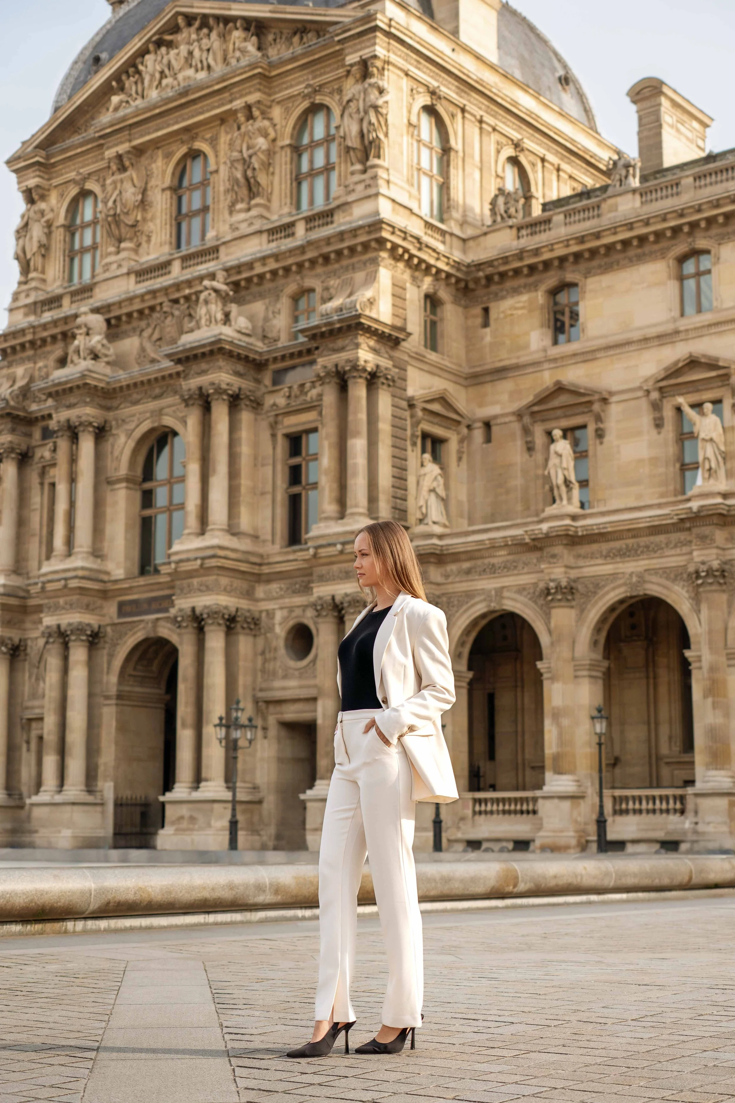 A woman in a white blazer and pants standing in front of an ornate historical building with statues and detailed architecture.