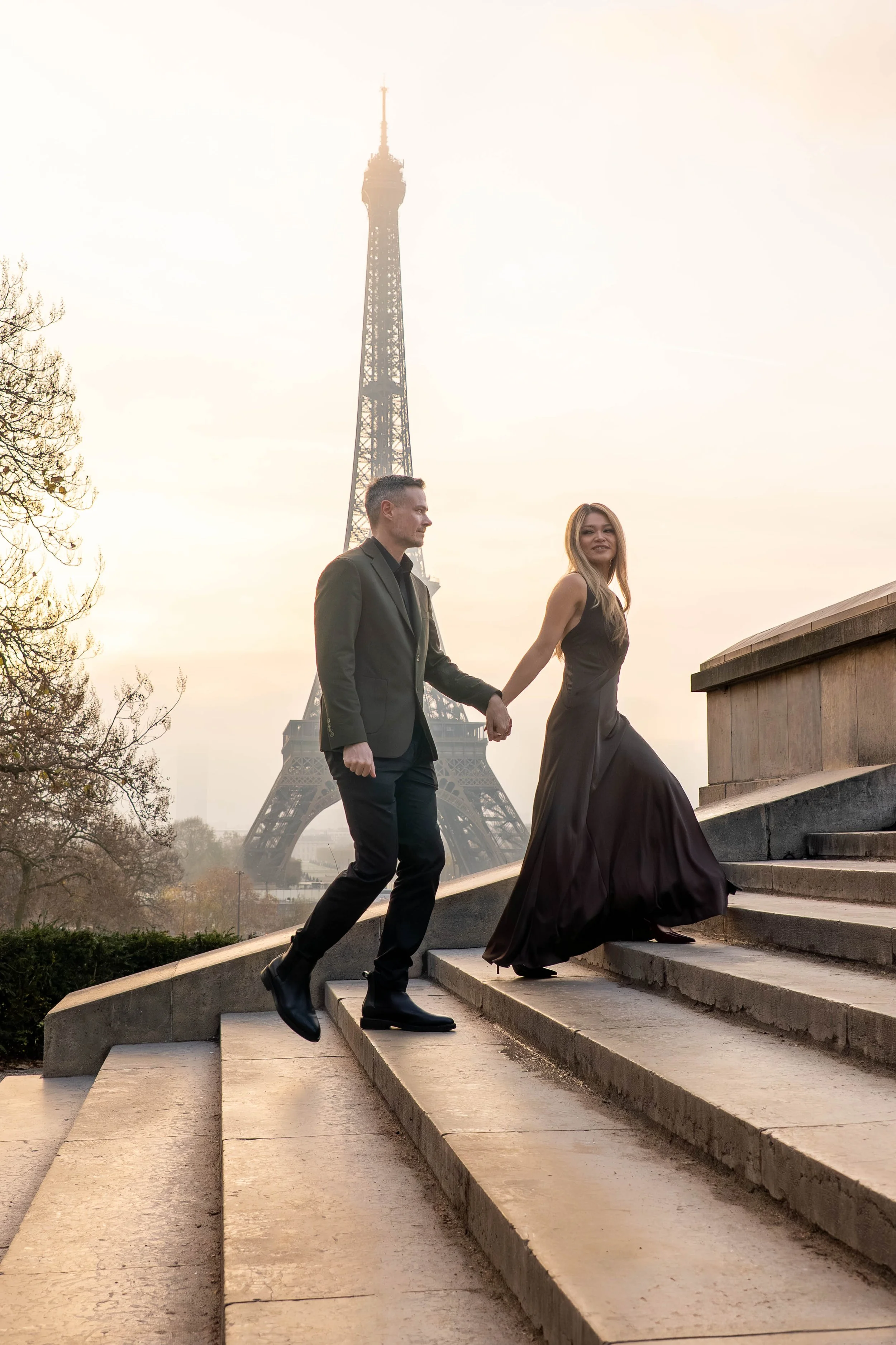 A couple holding hands and ascending stairs with the Eiffel Tower in the background during sunset. The woman is wearing a long black dress and the man is in a dark suit.