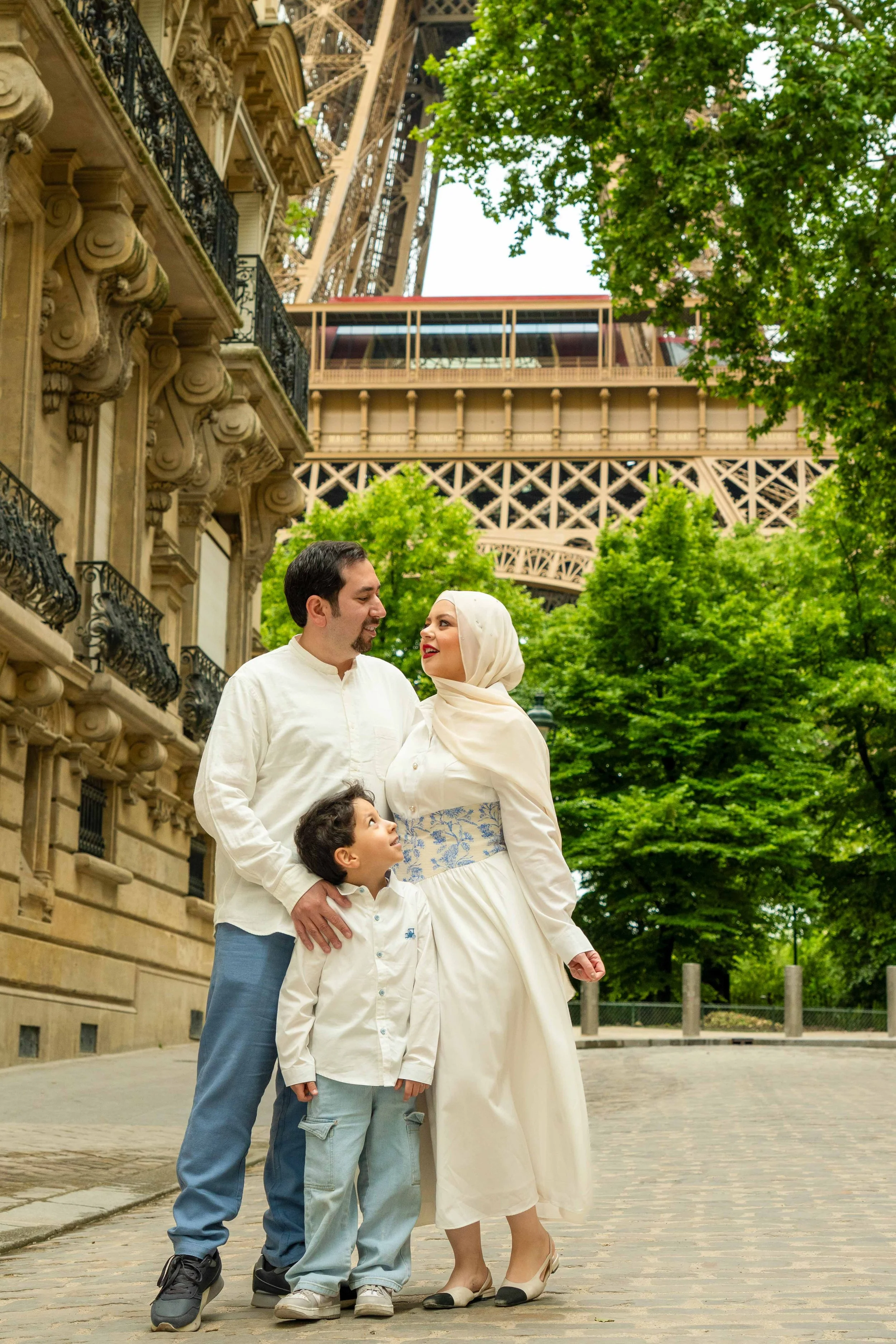 A family of three smiling and looking at each other outdoors near the Eiffel Tower in Paris, France, with green trees and historic buildings in the background.