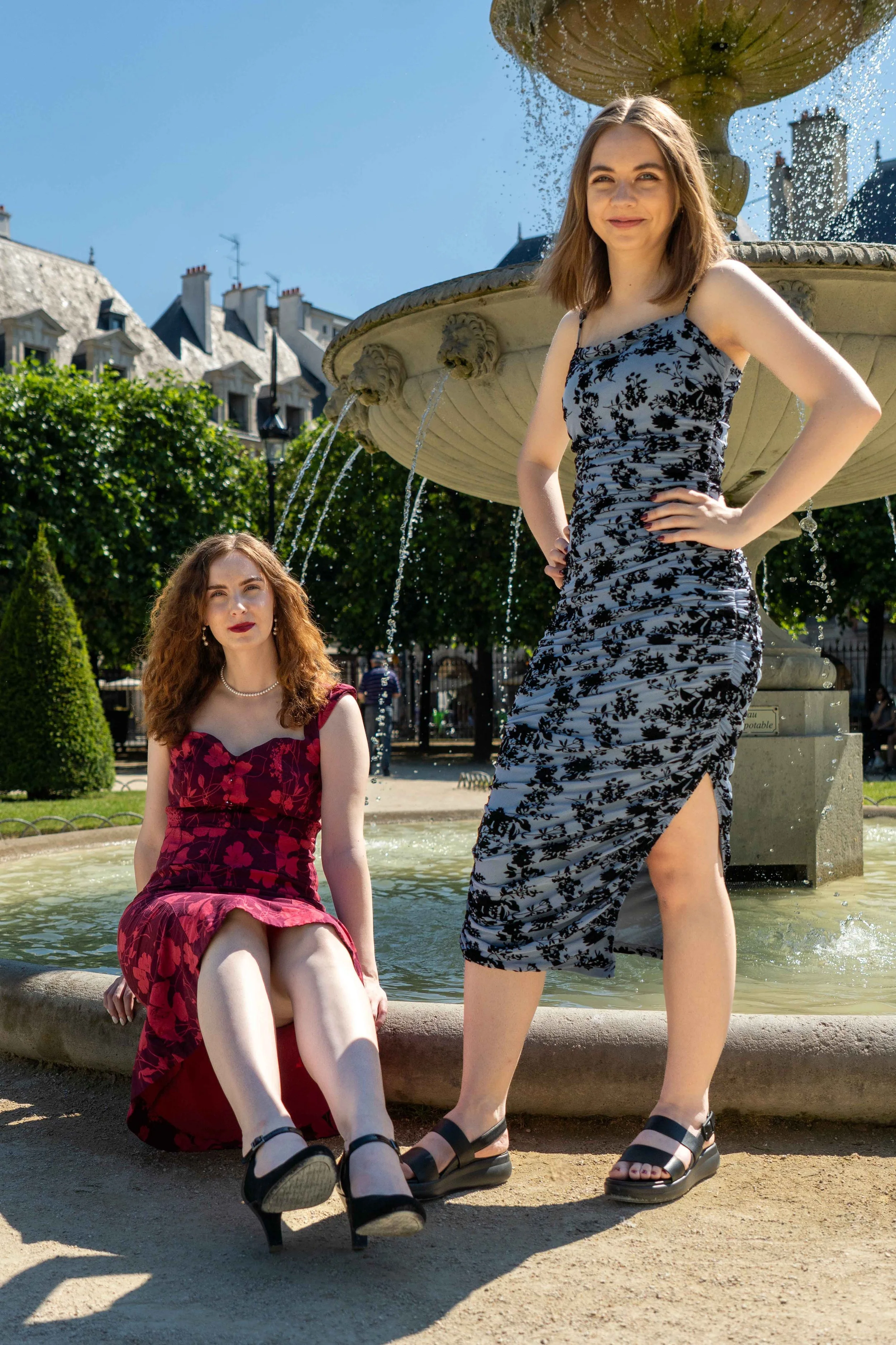 Two young women in stylish dresses posing in front of a fountain outdoors on a sunny day.