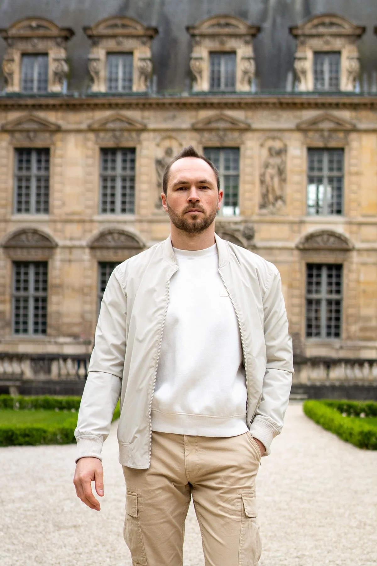 A young man with a beard and short dark hair standing outdoors in front of a historic stone building with ornate windows and sculptures, wearing a white jacket and beige pants.