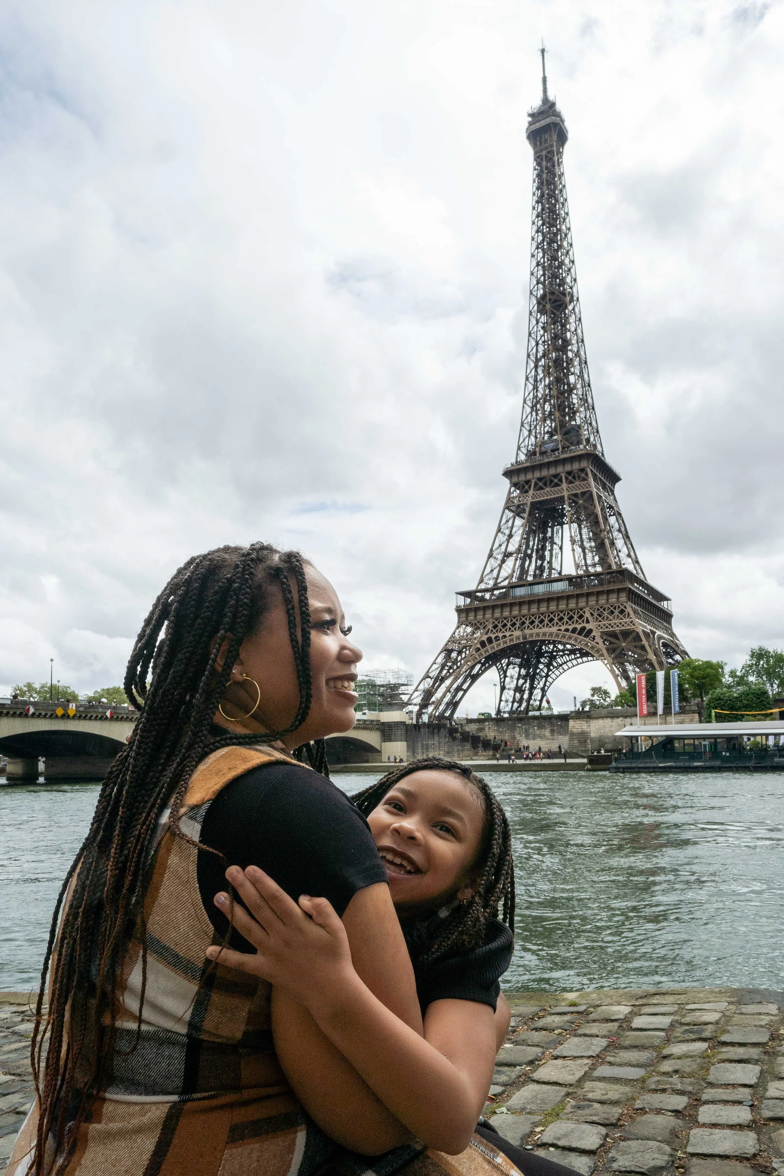 A woman and a young girl smiling and hugging near the Seine River with the Eiffel Tower in the background under a cloudy sky.