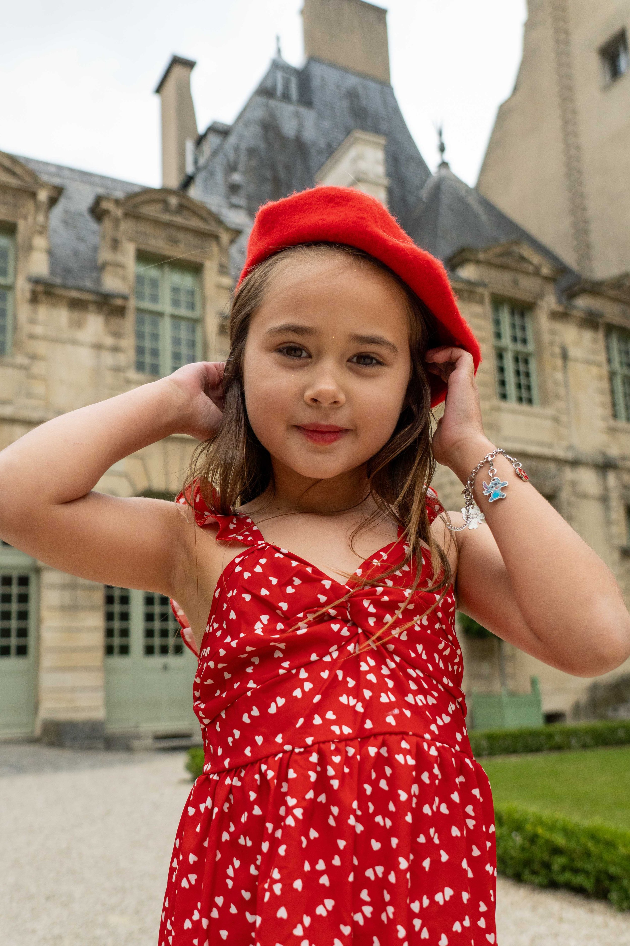 A young girl with brown hair wearing a red dress with white hearts and a red beret, standing outdoors in front of a historic stone building with turrets and trees.
