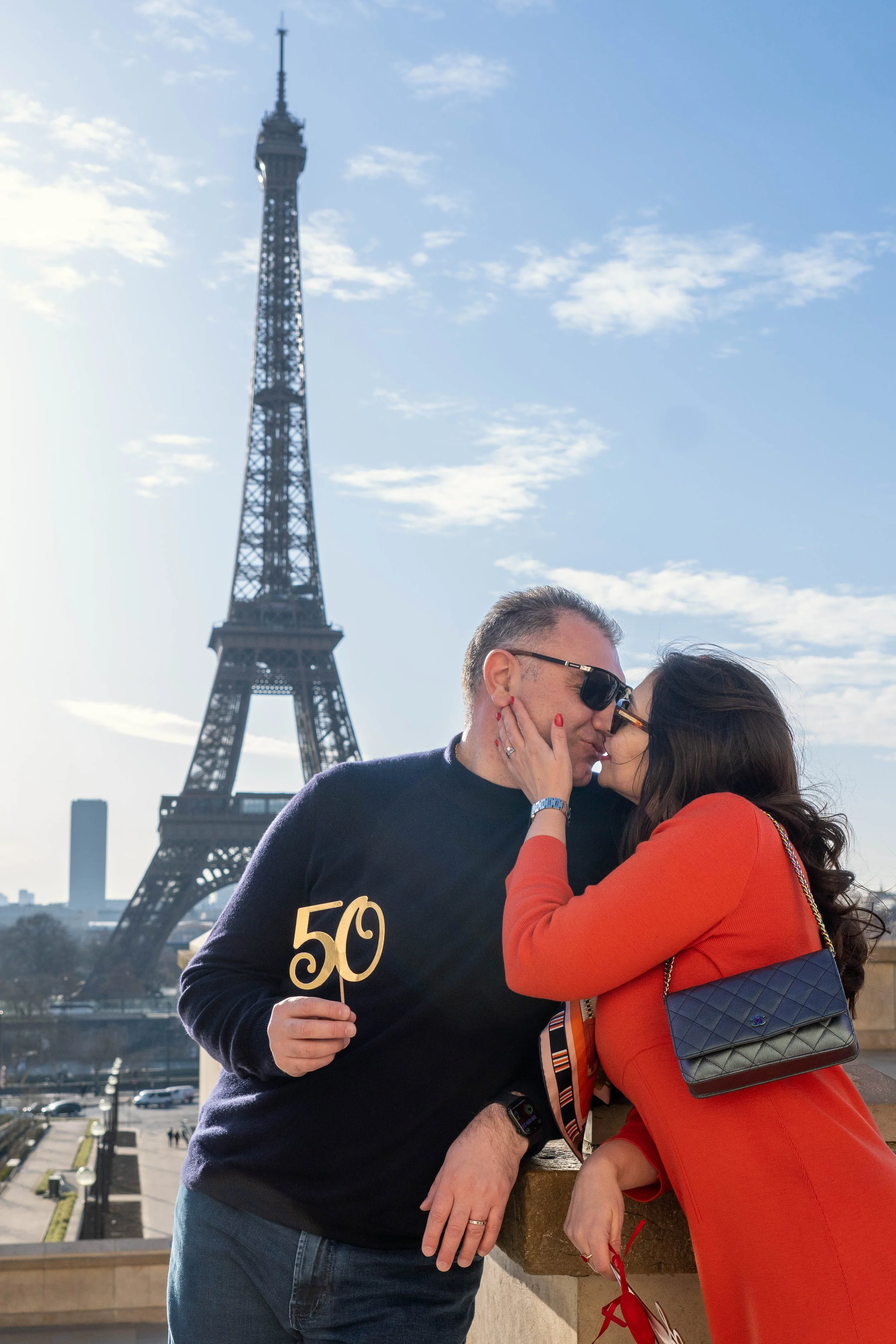 A couple sharing a kiss with the Eiffel Tower in the background. The man holds a sign with '50' on it. Both are dressed in casual attire, and the woman is carrying a quilted bag.