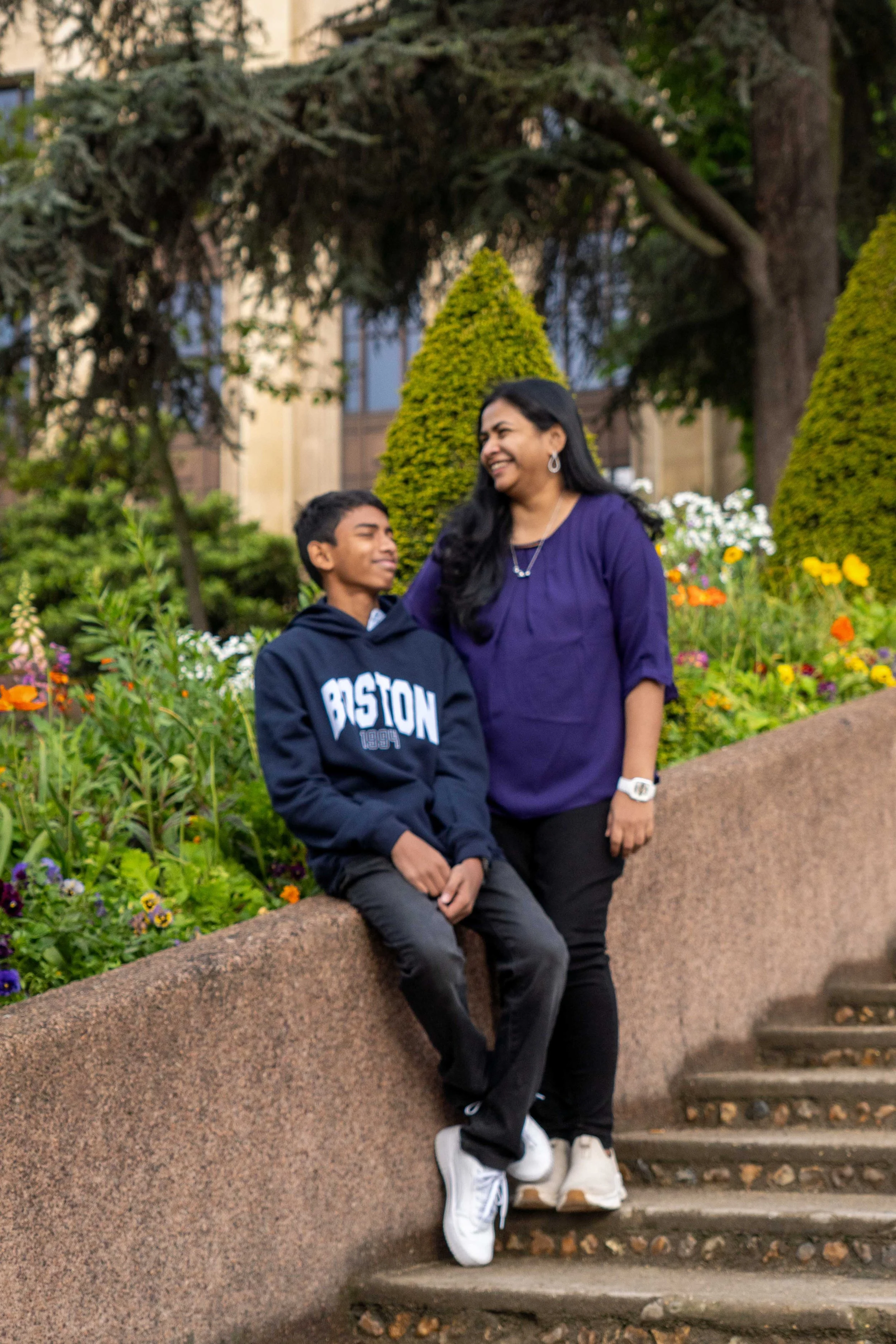 A woman and a teenage boy in a park with colorful flowers and greenery, smiling at each other.