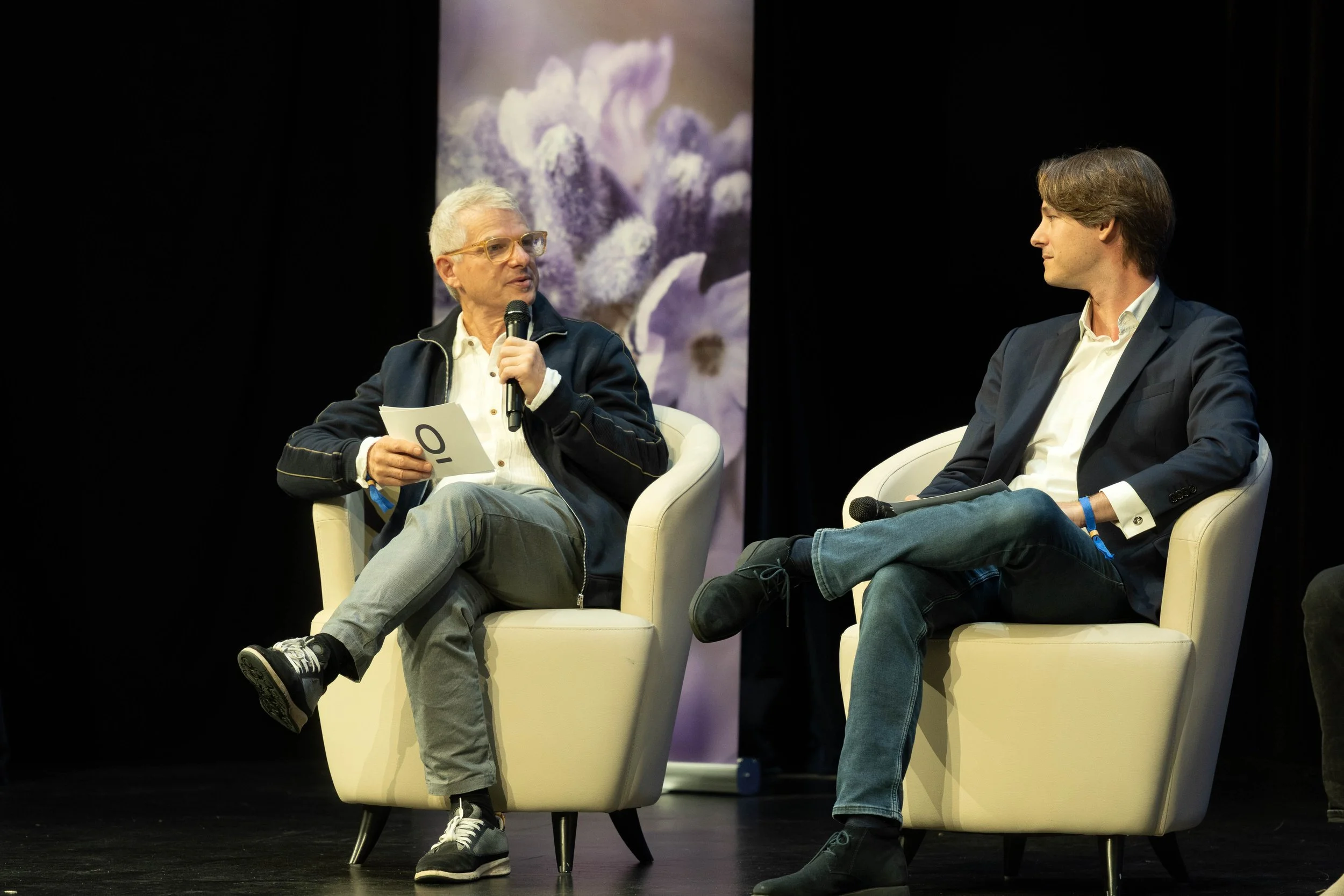 Two men in suits sitting on cream-colored chairs on stage during a discussion. The man on the left, with gray hair and glasses, is speaking into a microphone and holding notes. The man on the right, with brown hair, is listening attentively, also holding a microphone. There is a purple floral backdrop behind them.