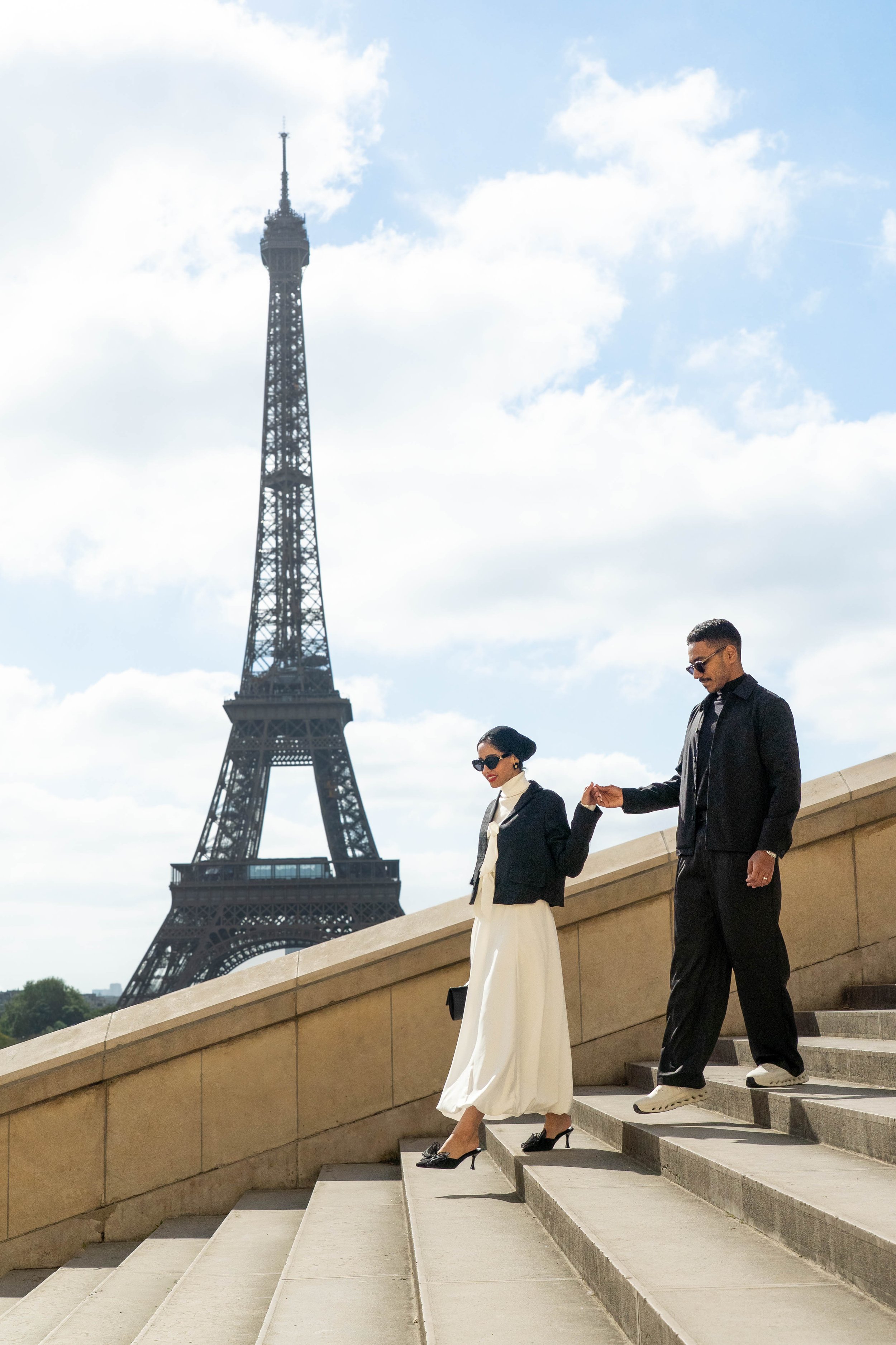 A man and woman holding hands while walking down stairs on the Eiffel Tower's viewing platform in Paris, France, with the Eiffel Tower in the background against a partly cloudy sky.
