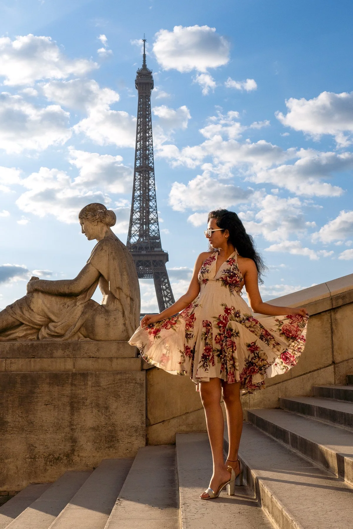 A woman in a floral dress and high heels standing on stairs near a stone statue with the Eiffel Tower in the background on a sunny day with clouds.