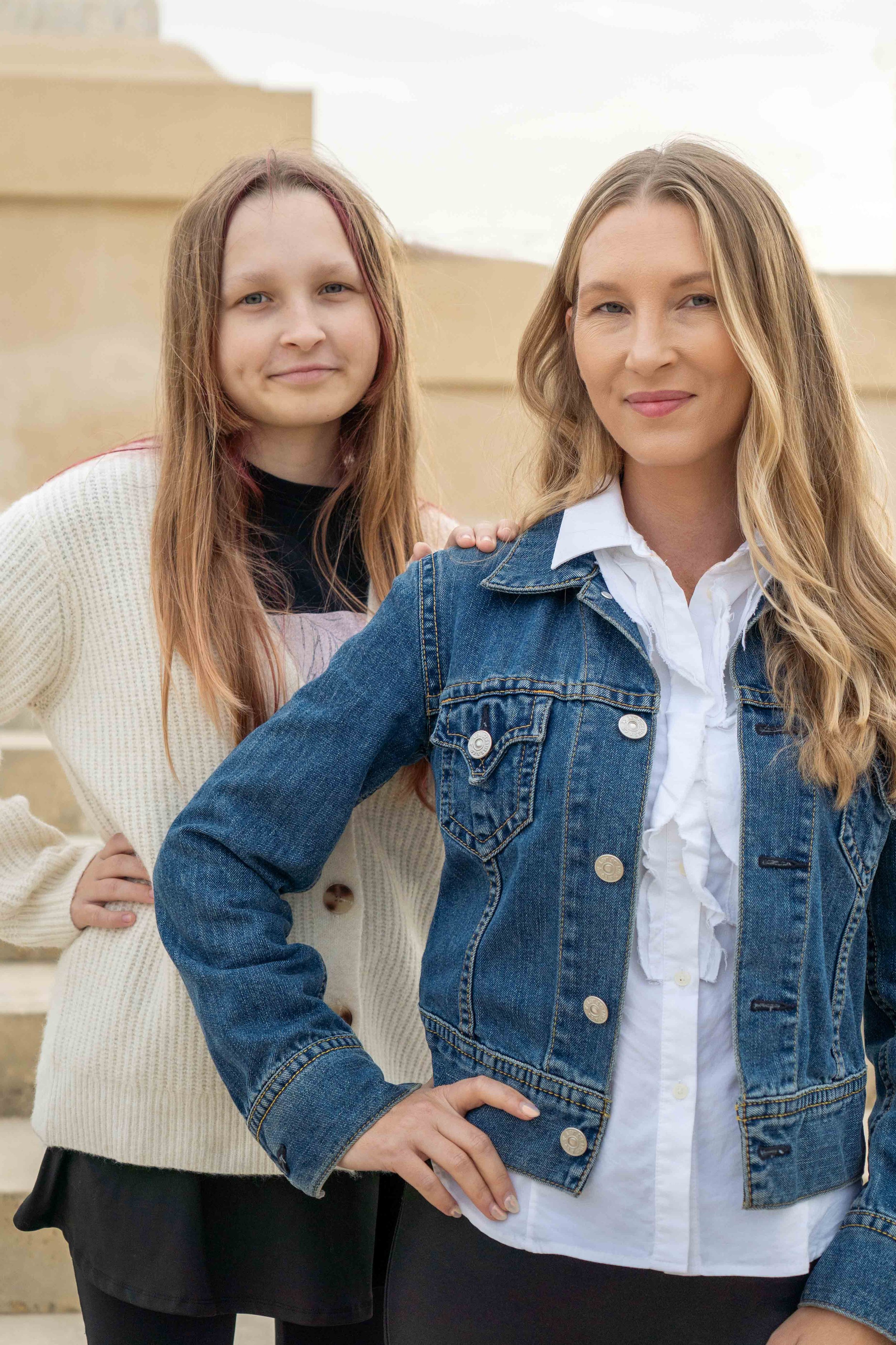 A woman and a girl standing outdoors, with the woman wearing a denim jacket and the girl wearing a white sweater, both looking at the camera.