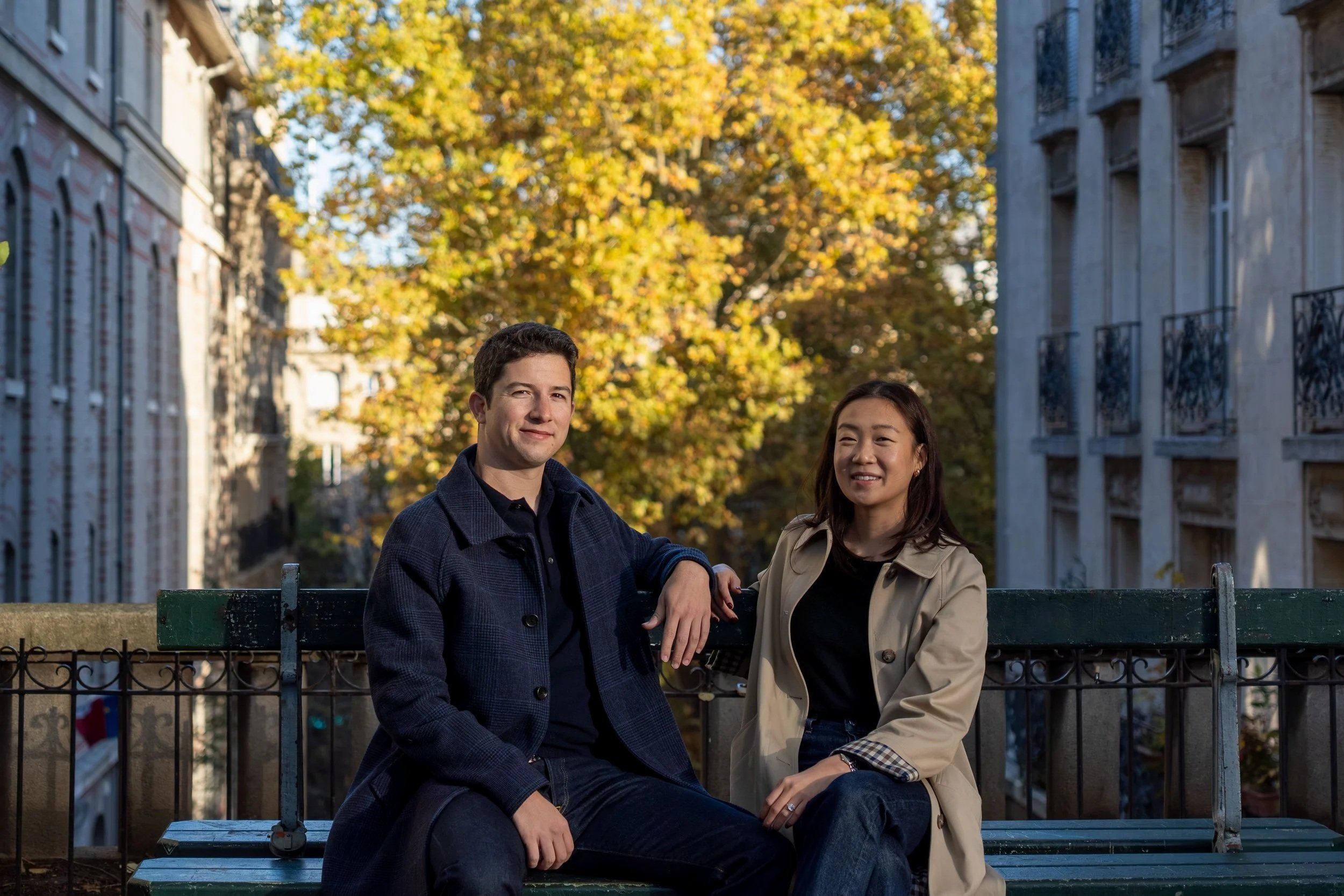 A man and a woman sitting on a park bench in an urban setting during fall, with trees with yellow and orange leaves in the background.