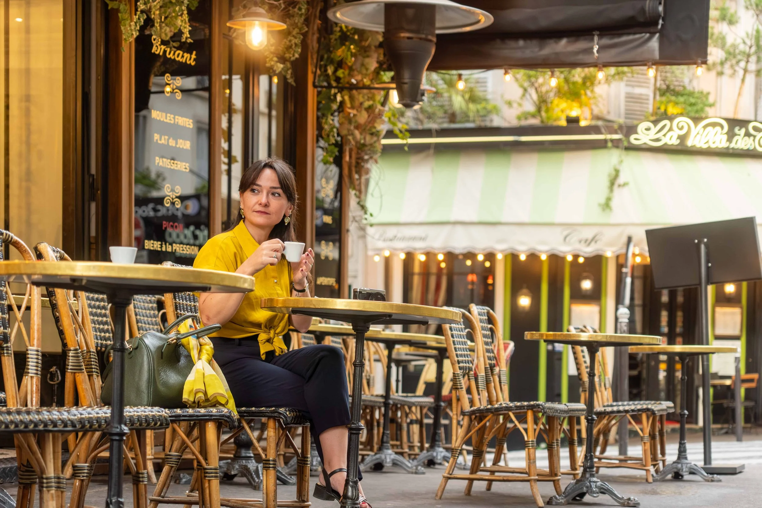 A woman with dark hair in a yellow blouse, sitting at an outdoor cafe table holding a white coffee cup, with empty tables and chairs around her.