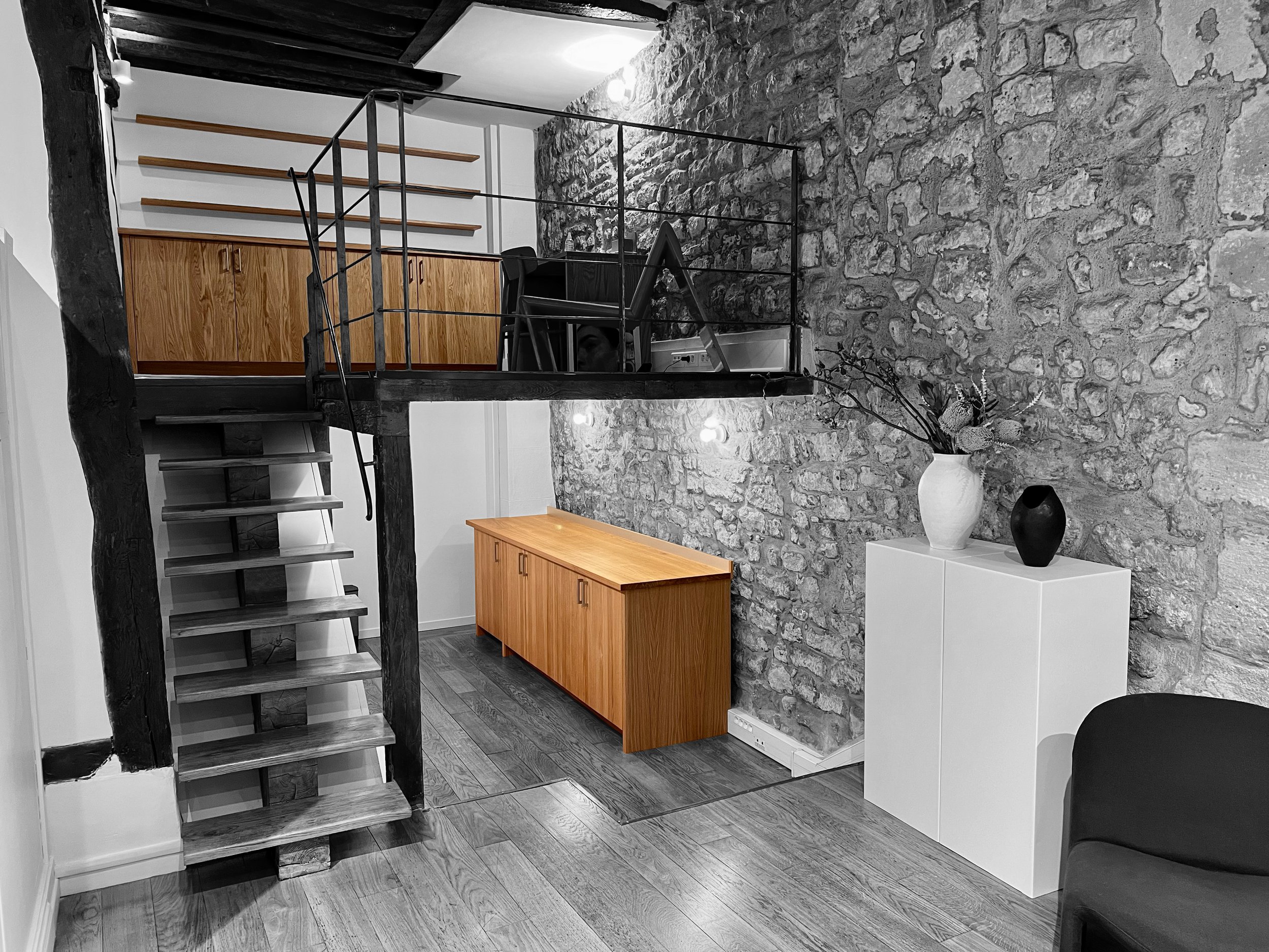Intérieur d'un loft avec mur en pierre, escalier en bois menant à une mezzanine avec un bureau, meuble en bois sous la mezzanine, deux vases noirs et un blanc avec des fleurs sur un meuble blanc, sol en bois.