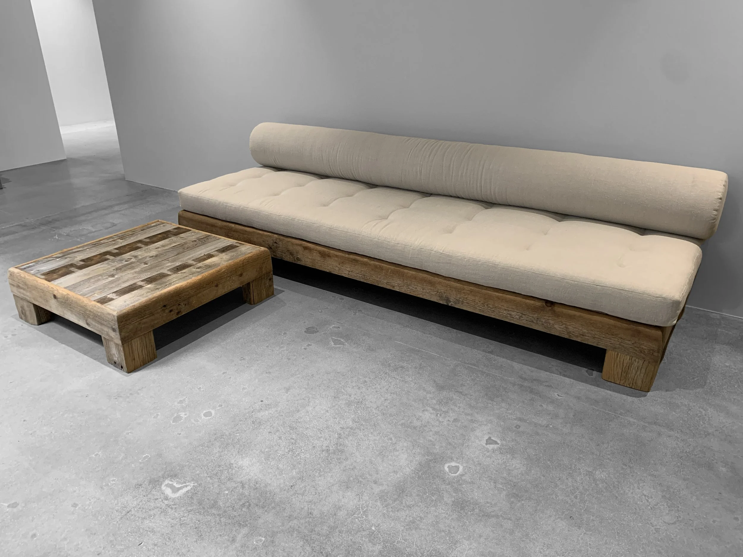 Canapé beige avec un cadre en bois clair et un coussin de dossier en bois, placé à côté d'une table basse en bois rustic, dans une pièce au sol en béton.