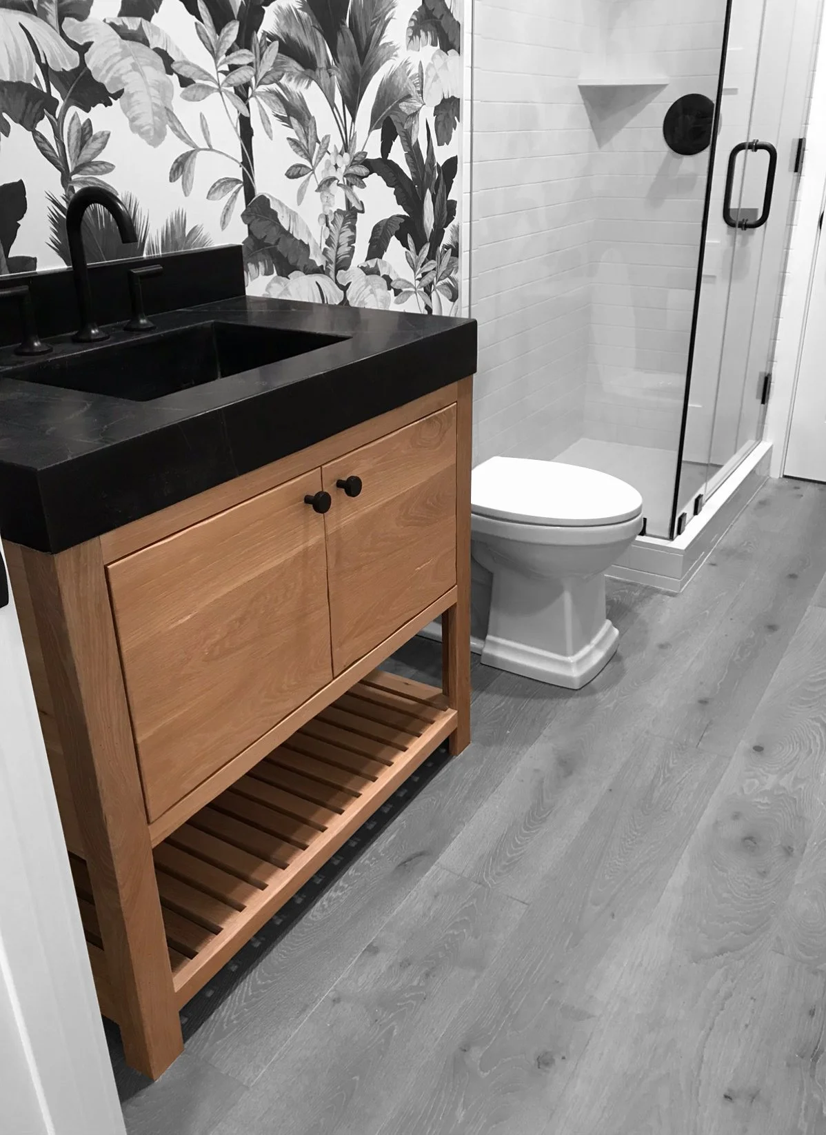 Salle de bain moderne avec lavabo en pierre noire, meuble en bois clair, mur de papier peint avec motif de feuilles, toilette blanche, douche en verre avec porte noire, plancher en bois clair.