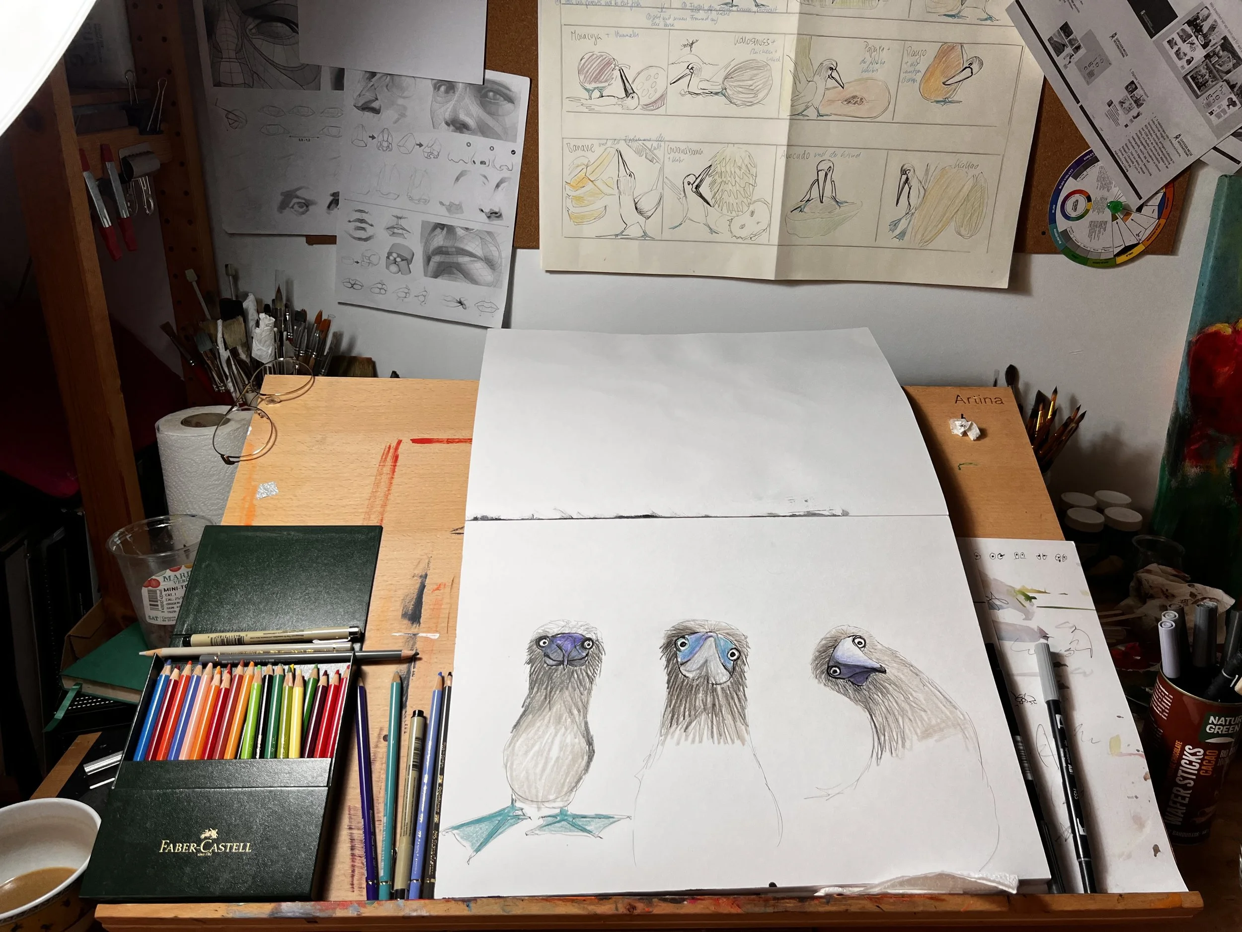 Zeichnungen und Skizzen von Vogelporträts auf einem Papier, umgeben von Buntstiften, Pinseln und Zeichenwerkzeugen auf einem Tisch in einem Kunststudio.