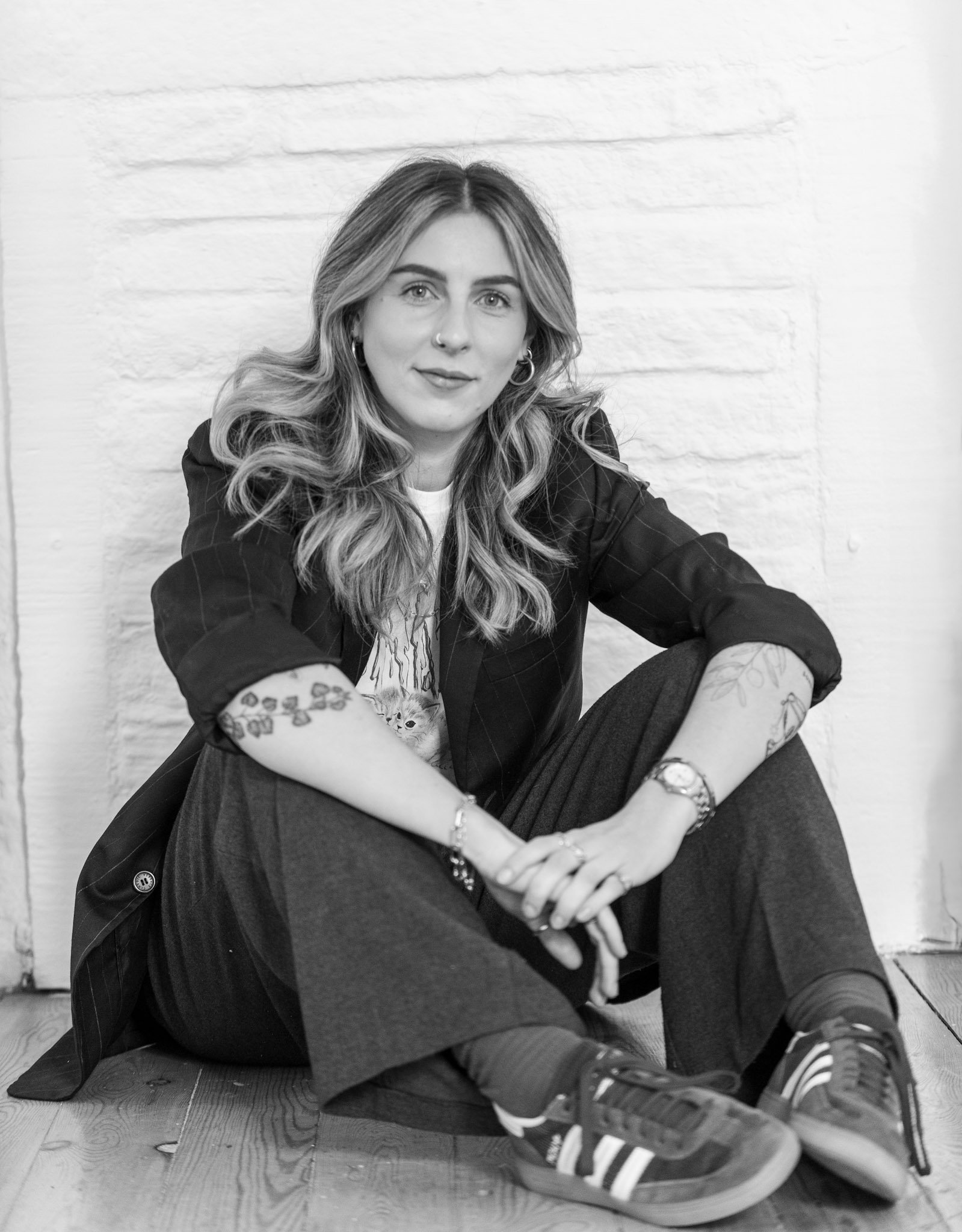 A woman with wavy hair, tattoos on her arms, wearing a jacket, shirt, pants, and sneakers, sitting on the floor against a white brick wall.