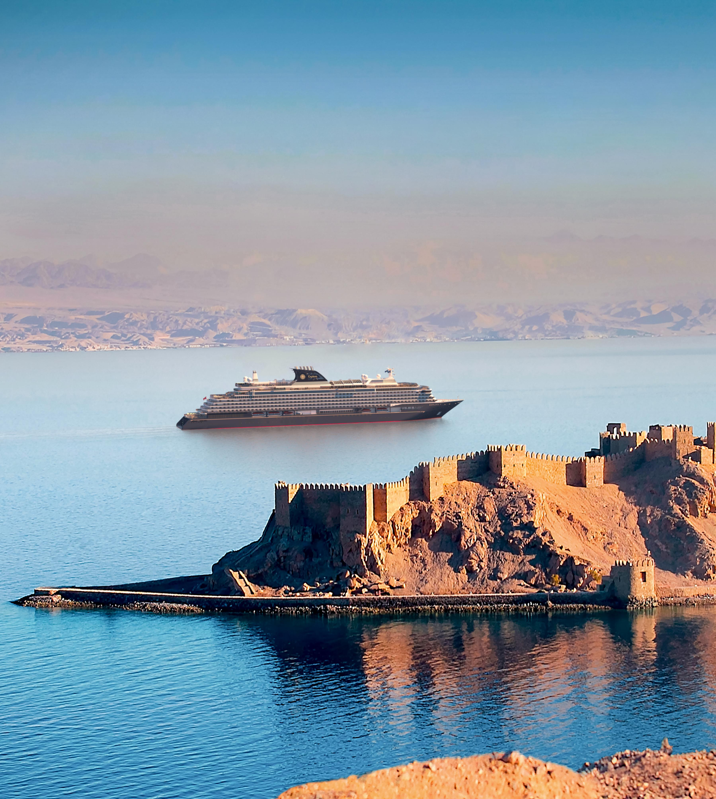 A large cruise ship sailing on a calm blue body of water near an island with historical castle walls and towers, with mountains in the background.