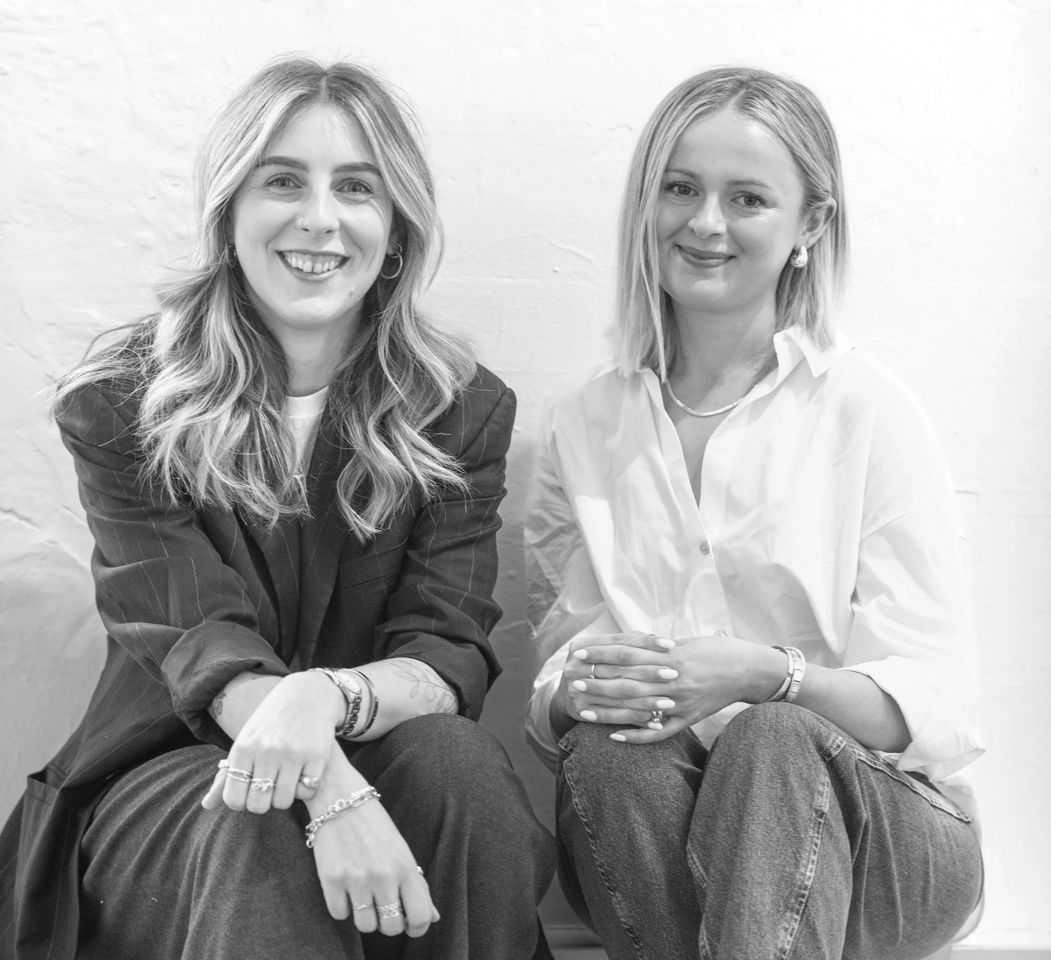 Two women sitting against a white brick wall, smiling for the camera.