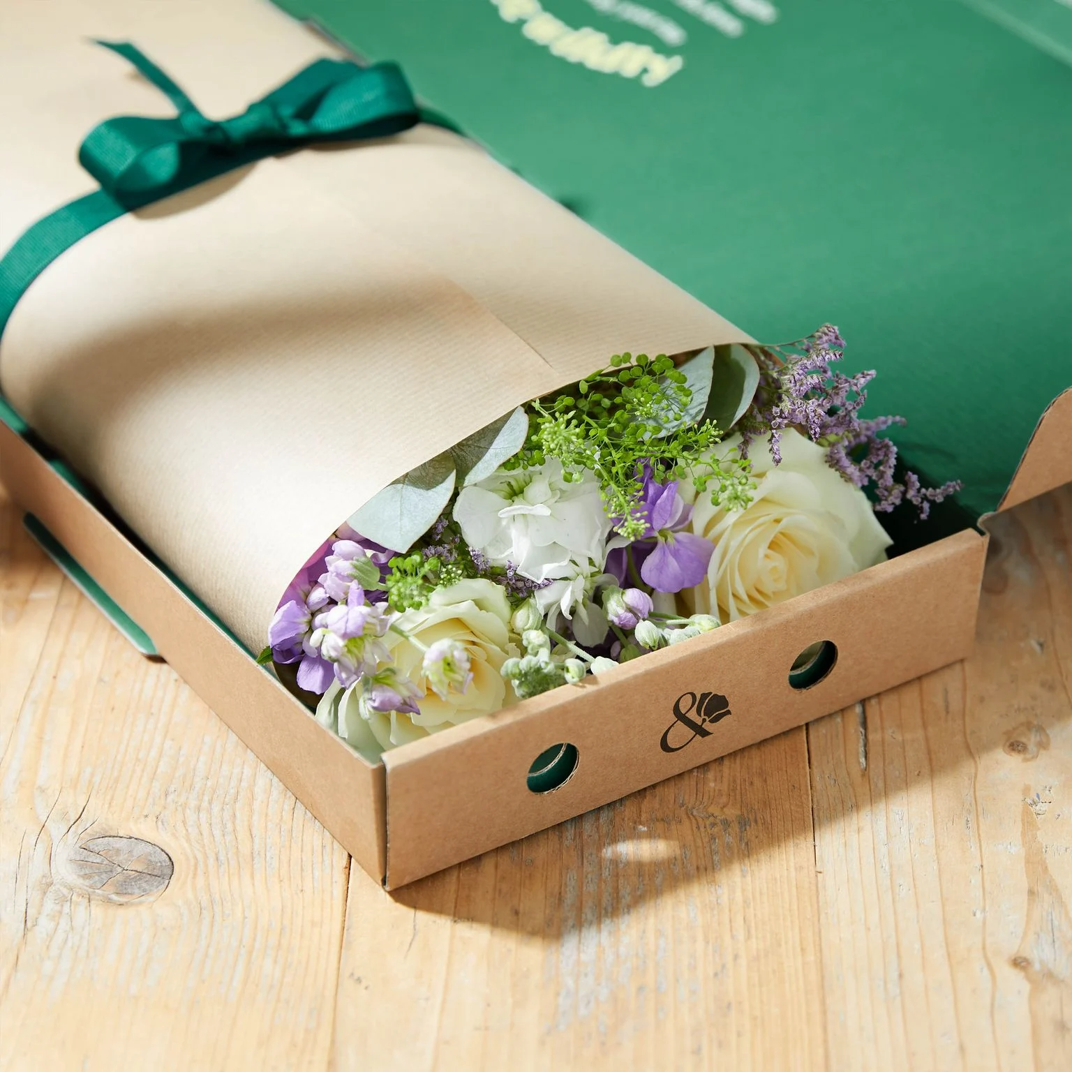 A flower bouquet with white roses and purple flowers in a cardboard box on a wooden surface.