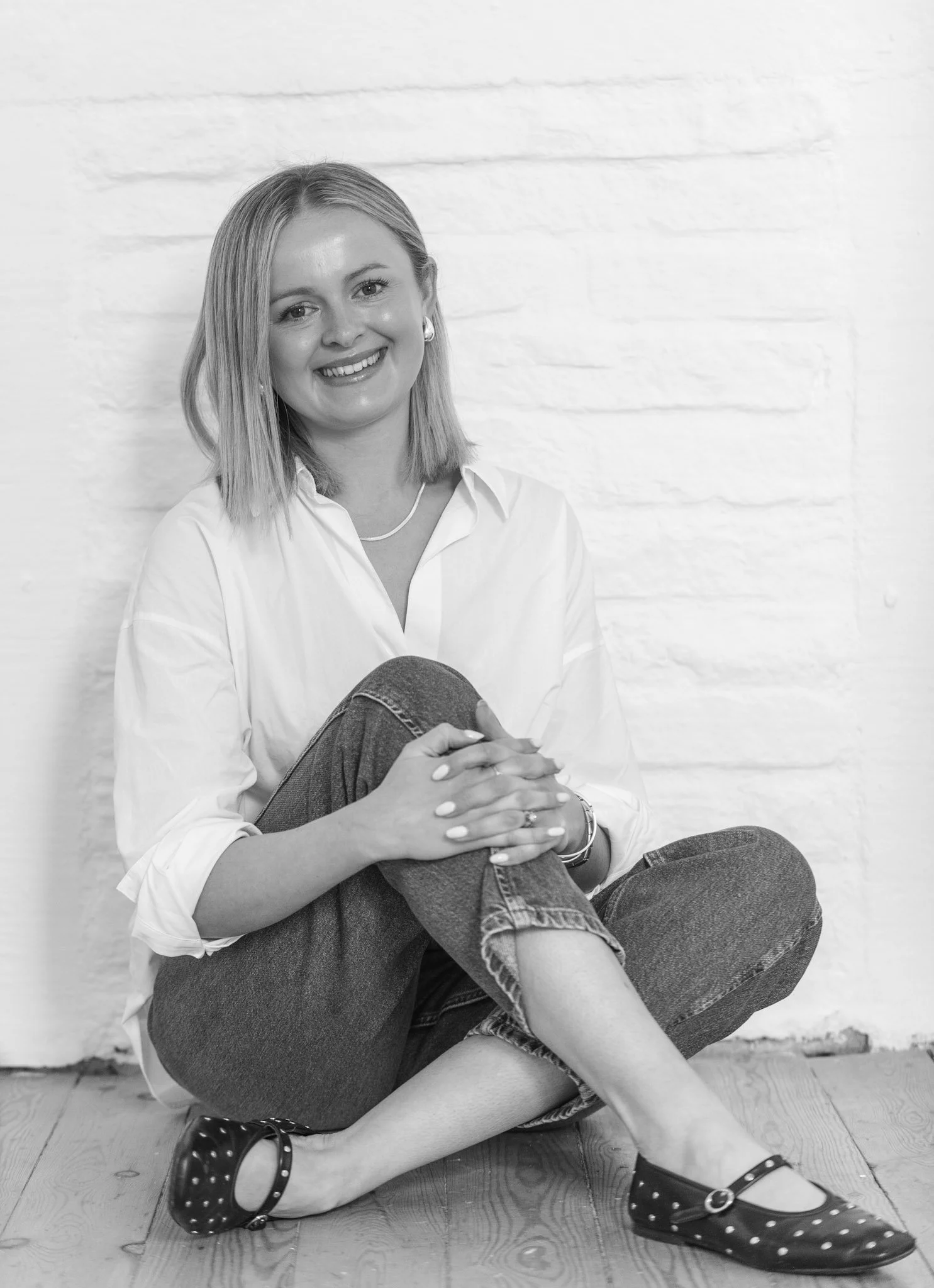 Black and white photo of a smiling woman sitting on hardwood floor against a white brick wall, wearing a white shirt, jeans, and black shoes with white polka dots.