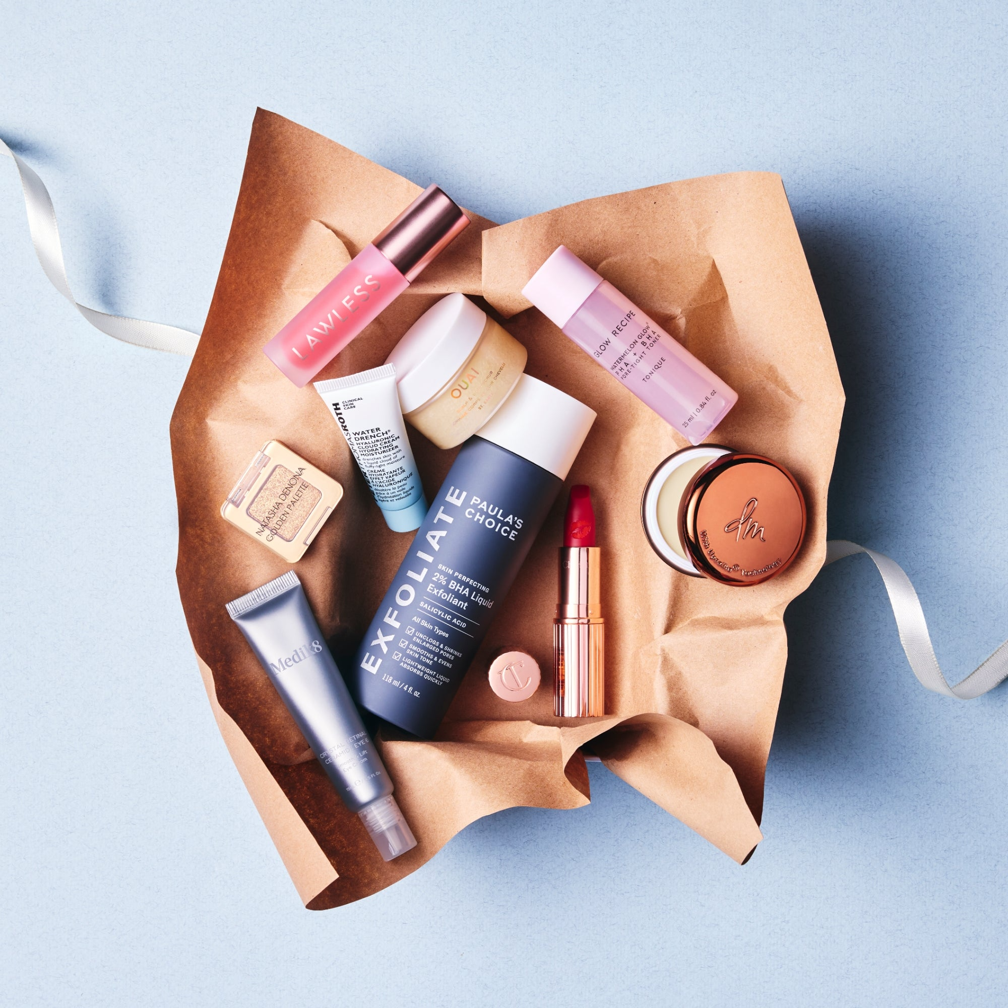 Collection of skincare and beauty products including creams, a lipstick, and serums arranged on crumpled brown paper with a blue background.
