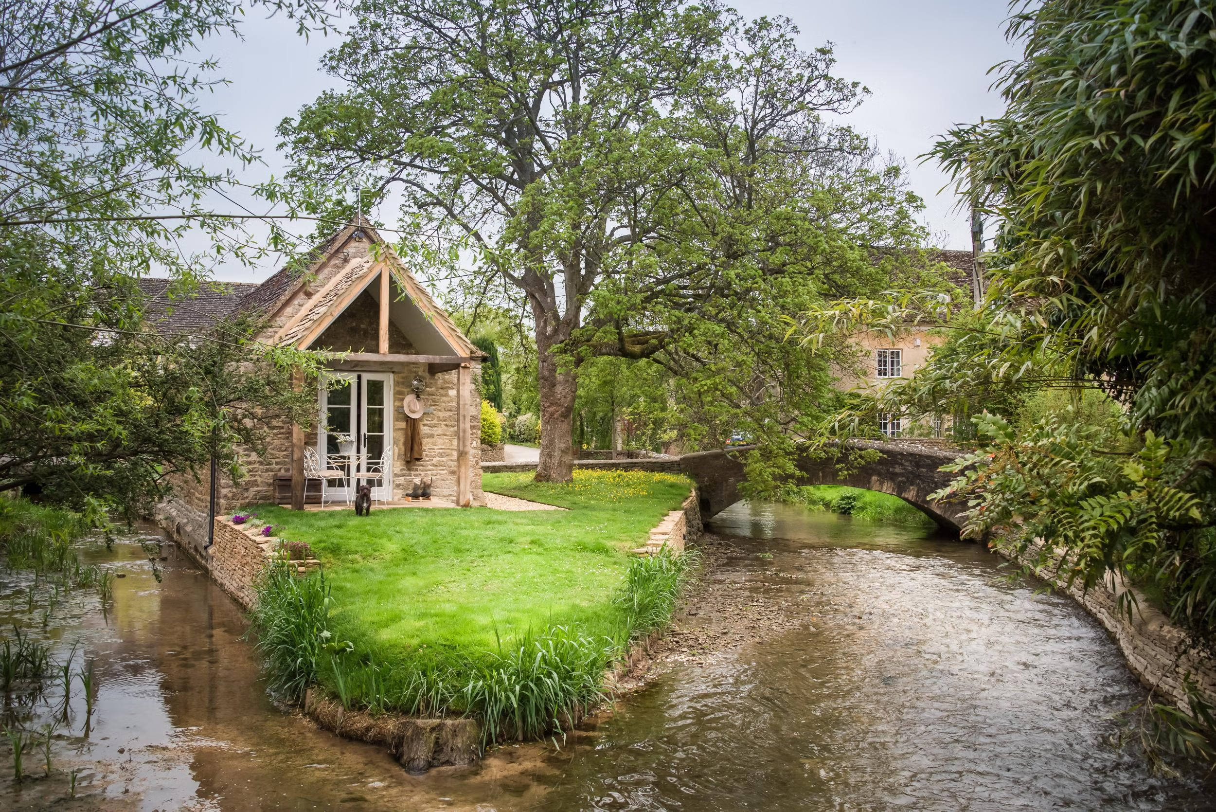 A small stone house with a porch, surrounded by greenery, located beside a flowing stream with a stone bridge in the background.