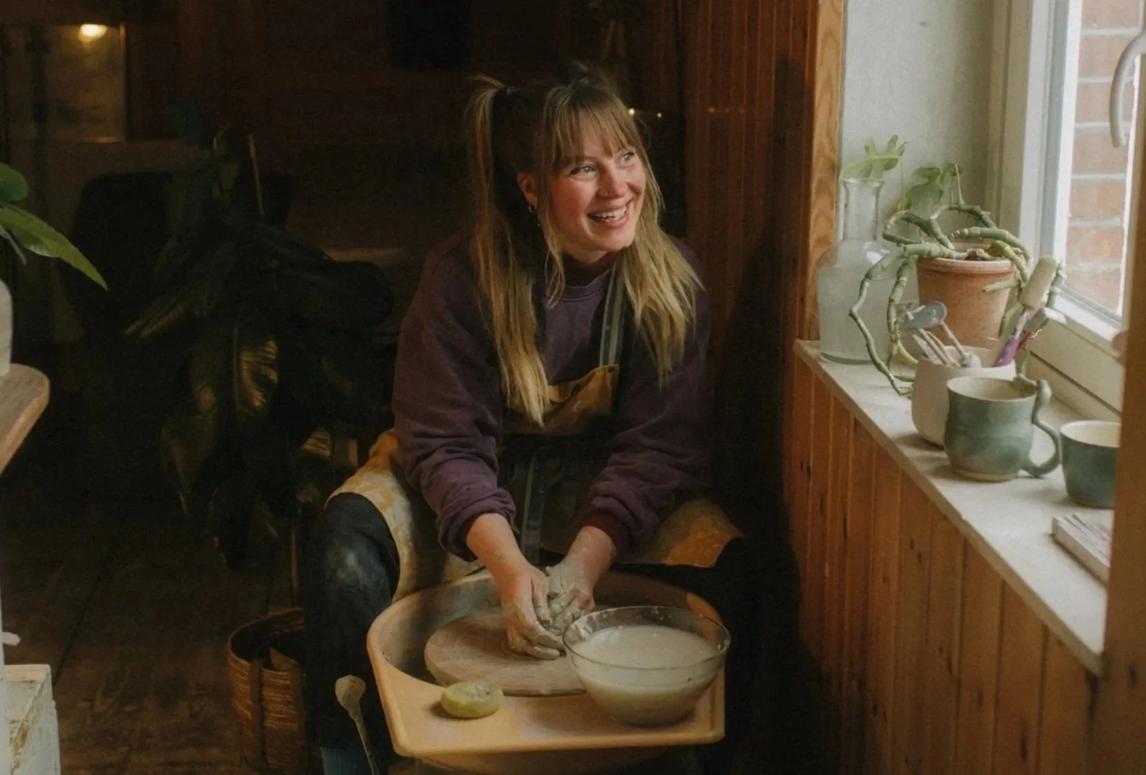 Eine lachende junge Frau beim Töpfern in einer gemütlichen, holzvertäfelten Küche, mit verschiedenen Tontöpfen auf der Fensterbank.