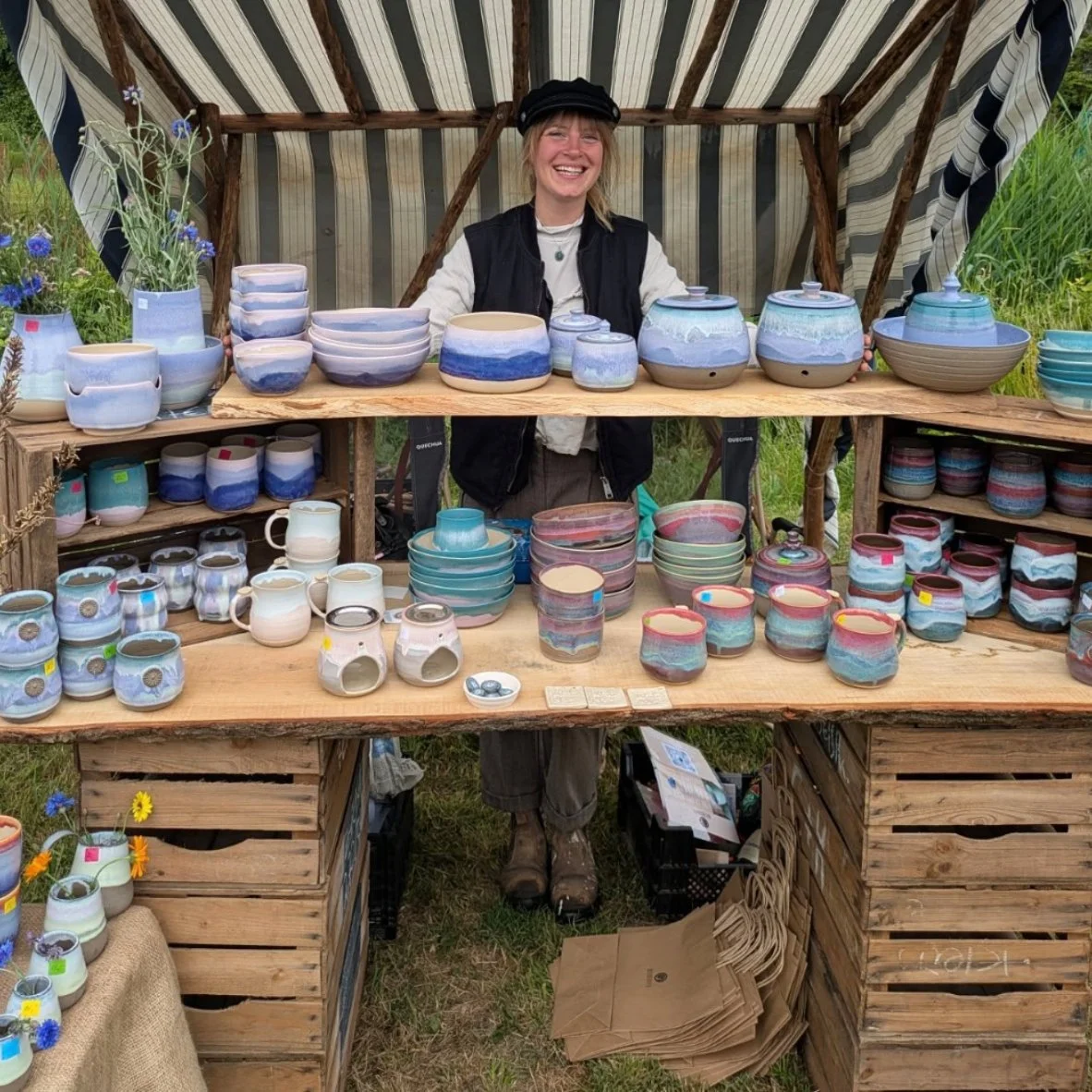 Lächelnde Frau steht hinter einem Tisch mit handgemachtem Keramikgeschirr, auf einem Markt im Freien.
