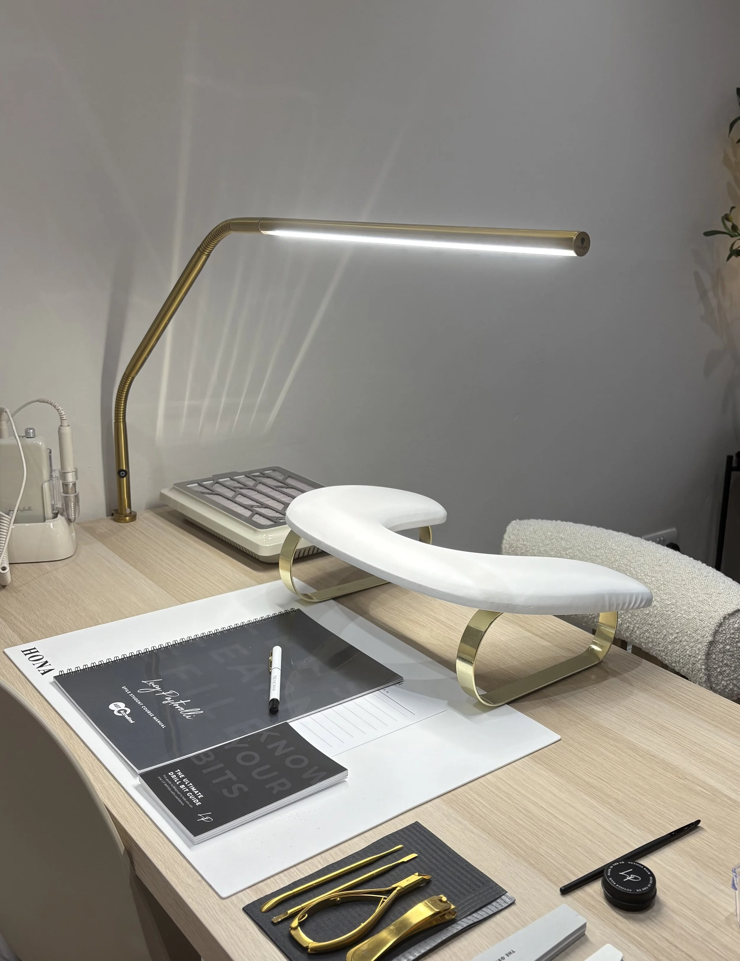 A modern, minimalist workspace featuring a wooden desk with a white and gold footrest, a spiral-bound notebook, a pen, and a pair of gold scissors. There is a sleek, gold desk lamp, some documents, a computer mouse, and a few electronic devices on the desk, with a gray and white wall in the background.