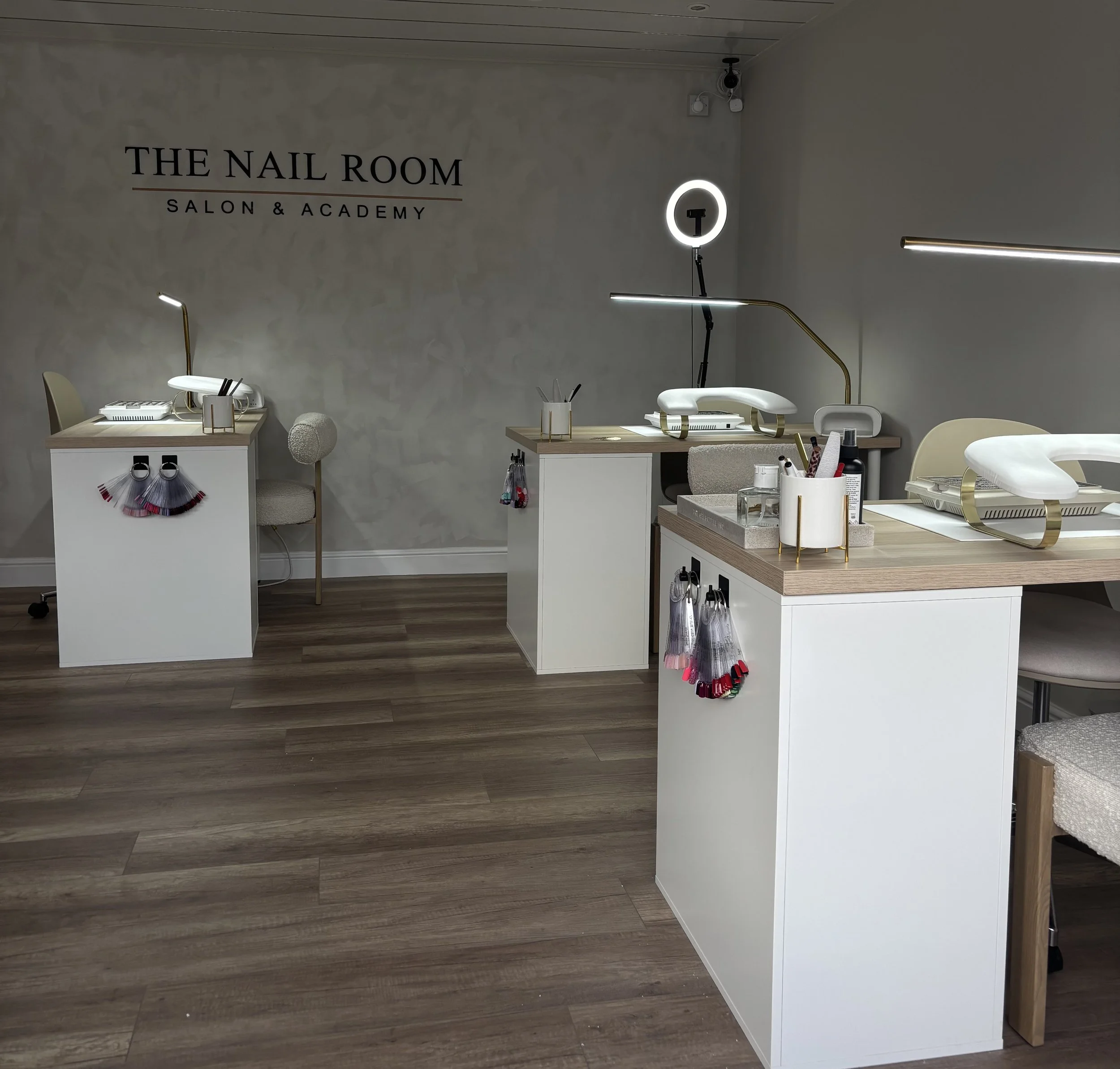 Interior of a nail salon with three manicure stations, white chairs, tools, and decor. The wall has the sign 'The Nail Room Salon & Academy.'