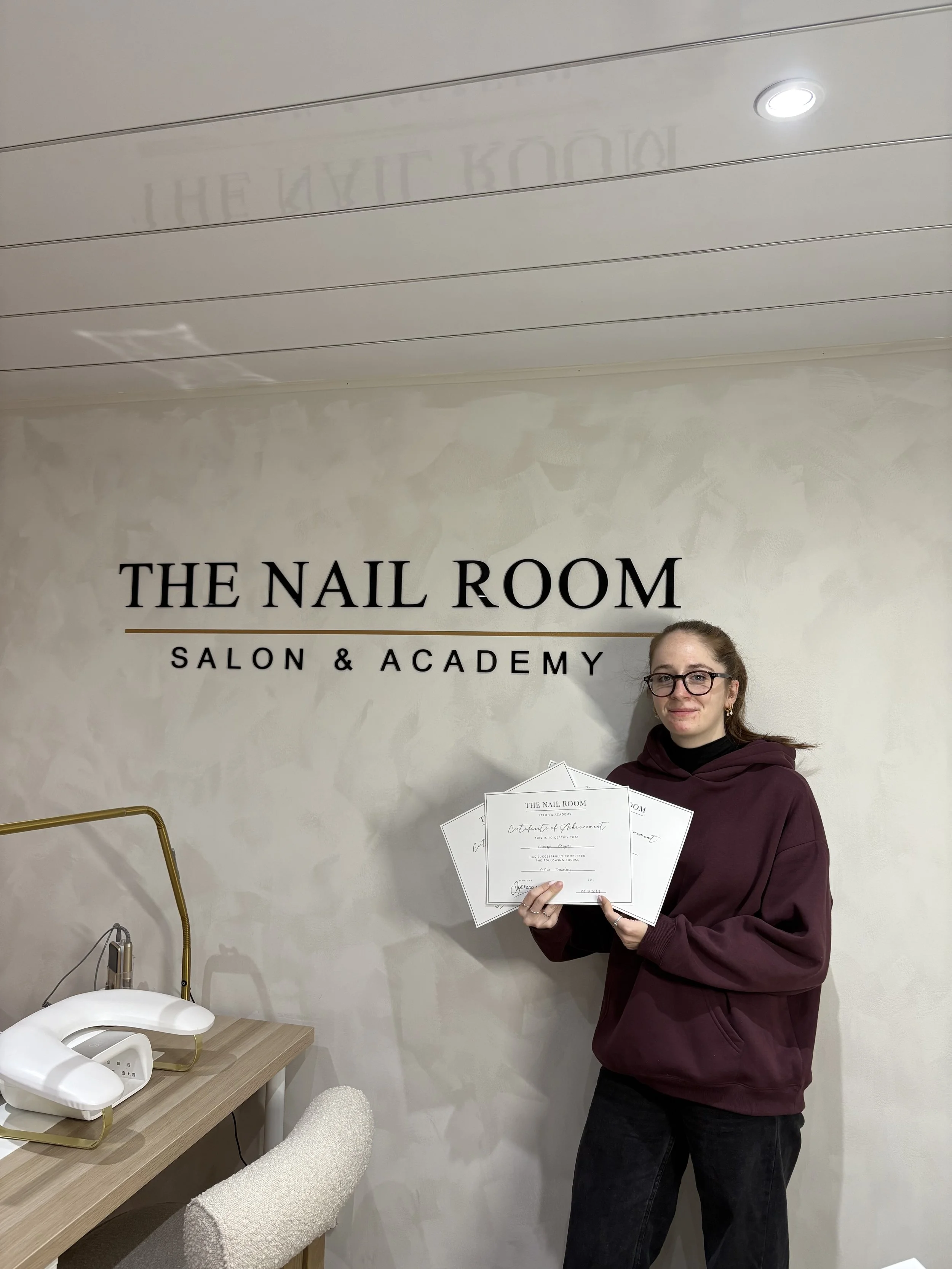 A young woman with glasses and a maroon hoodie standing in front of a wall with the sign 'The Nail Room Salon & Academy'. She is holding several certificates of achievement.