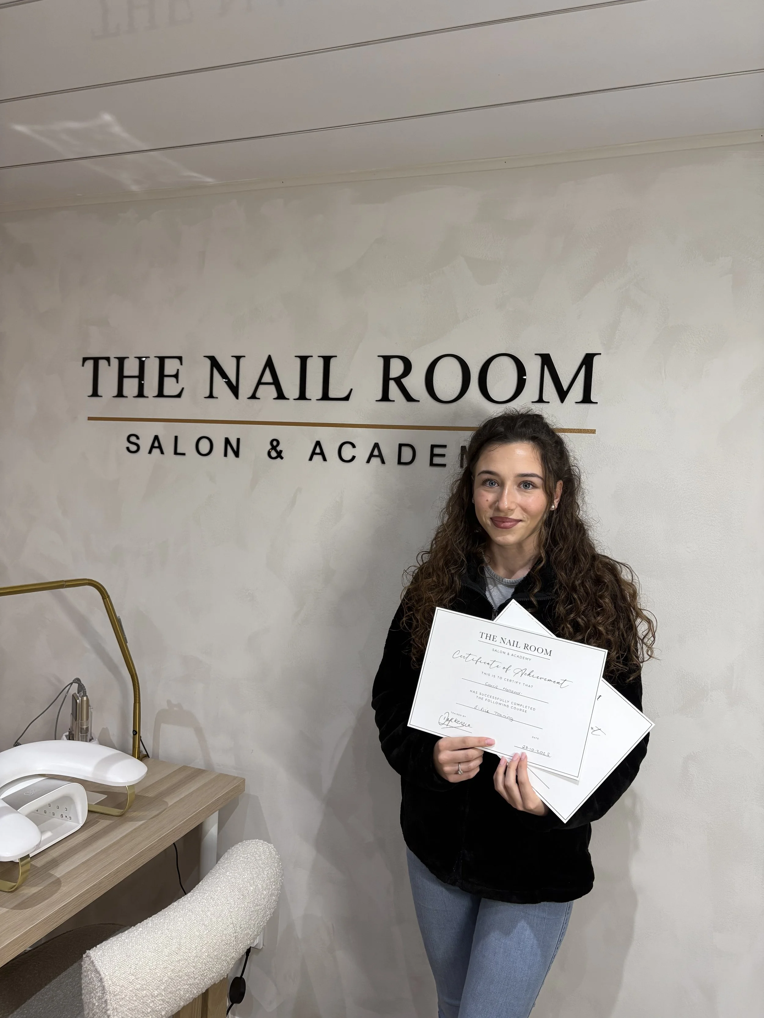 Young woman with curly hair holding certificates, standing in front of a wall with sign that reads "The Nail Room Salon & Academy."