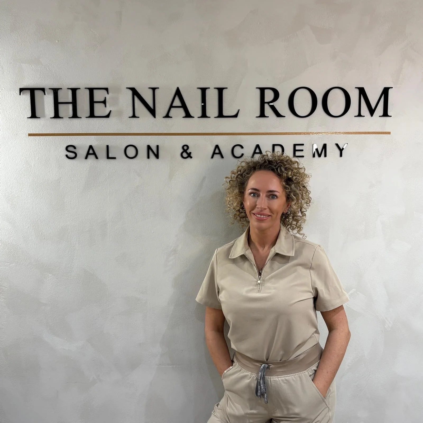 A woman with curly blonde hair standing in front of a sign that reads 'The Nail Room Salon & Academy' in black letters on a light-colored wall.