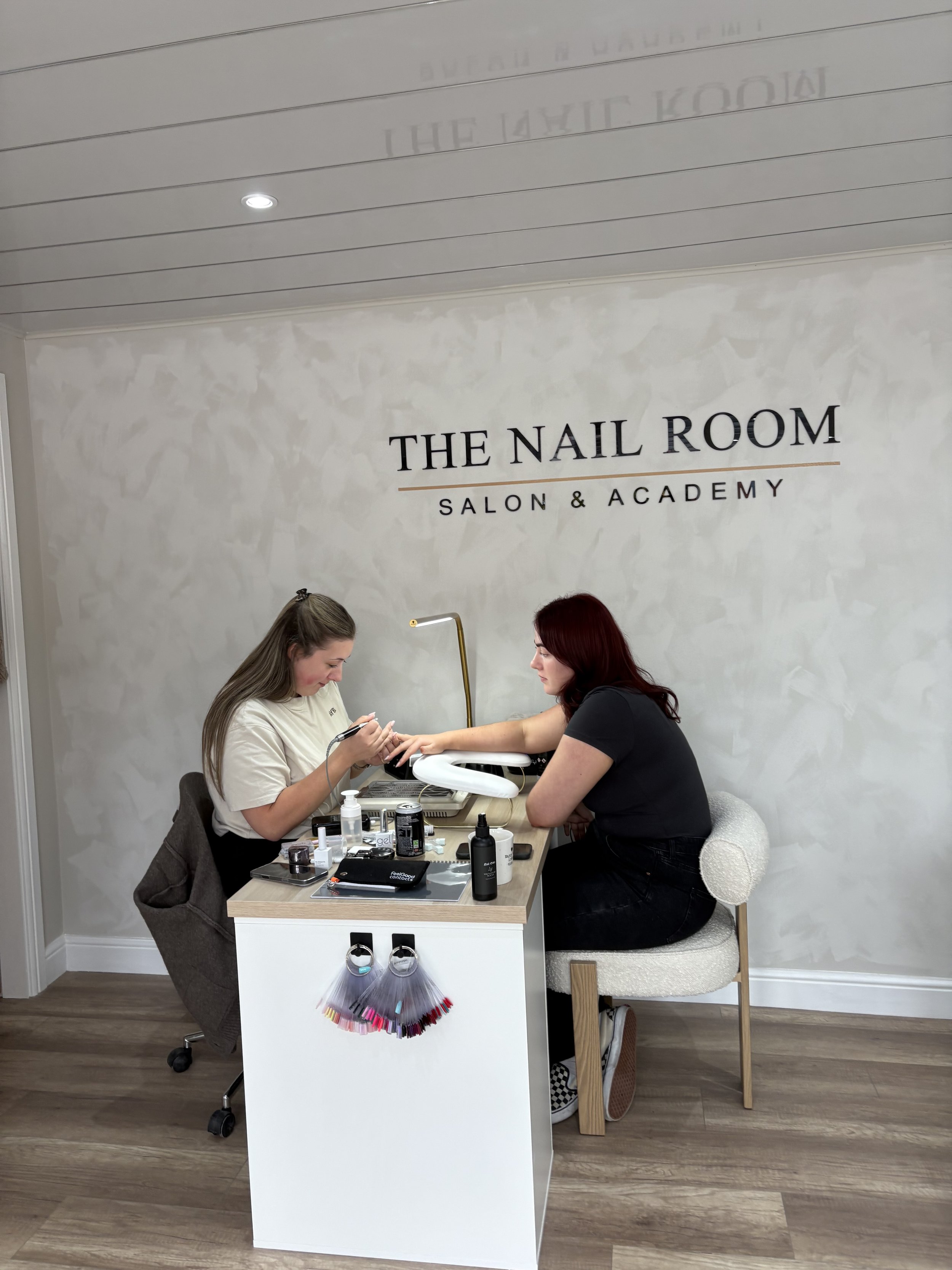 A woman training in gel nail application. The nail technician is focused on her training work, using tools on the woman's hands. The salon's wall has a sign that reads "The Nail Room, Salon & Academy."