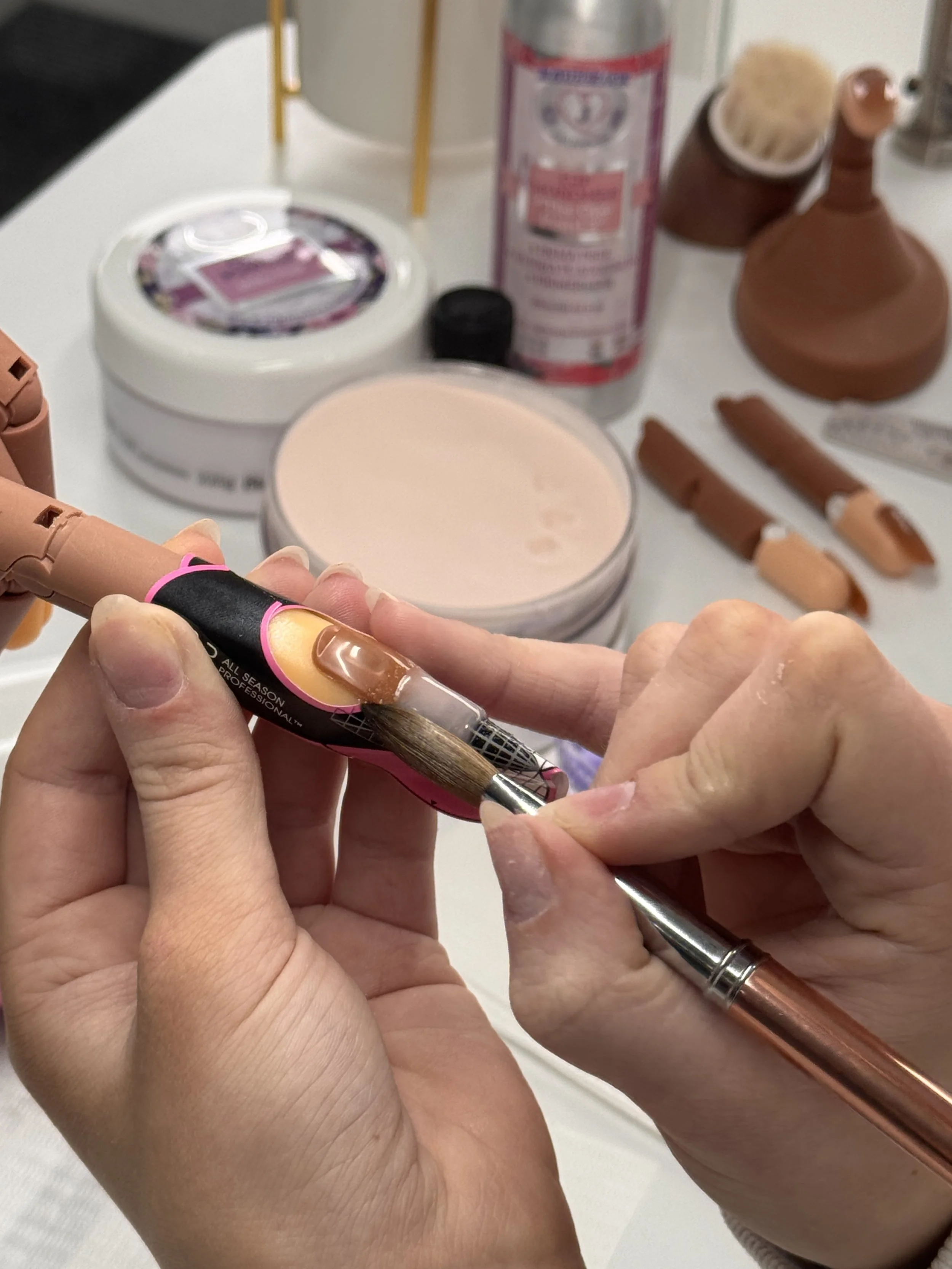 Close-up of a person applying nail polish with a small brush, with various manicure tools and products on a table in the background.