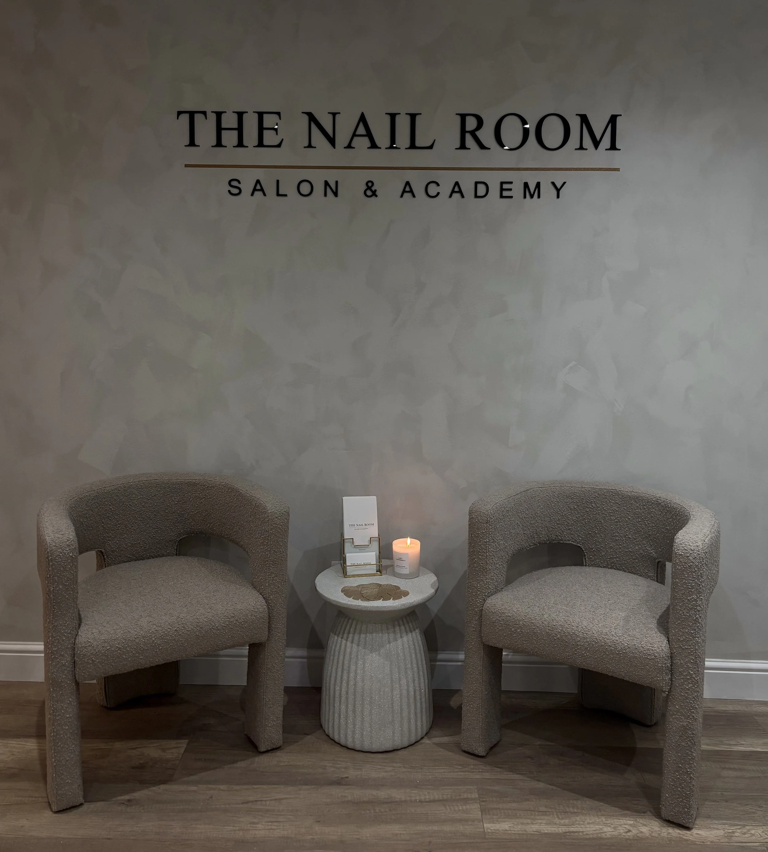Waiting area at The Nail Room Salon & Academy with two beige chairs, a small round textured table, a lit candle, a tray of nails, and a sign.